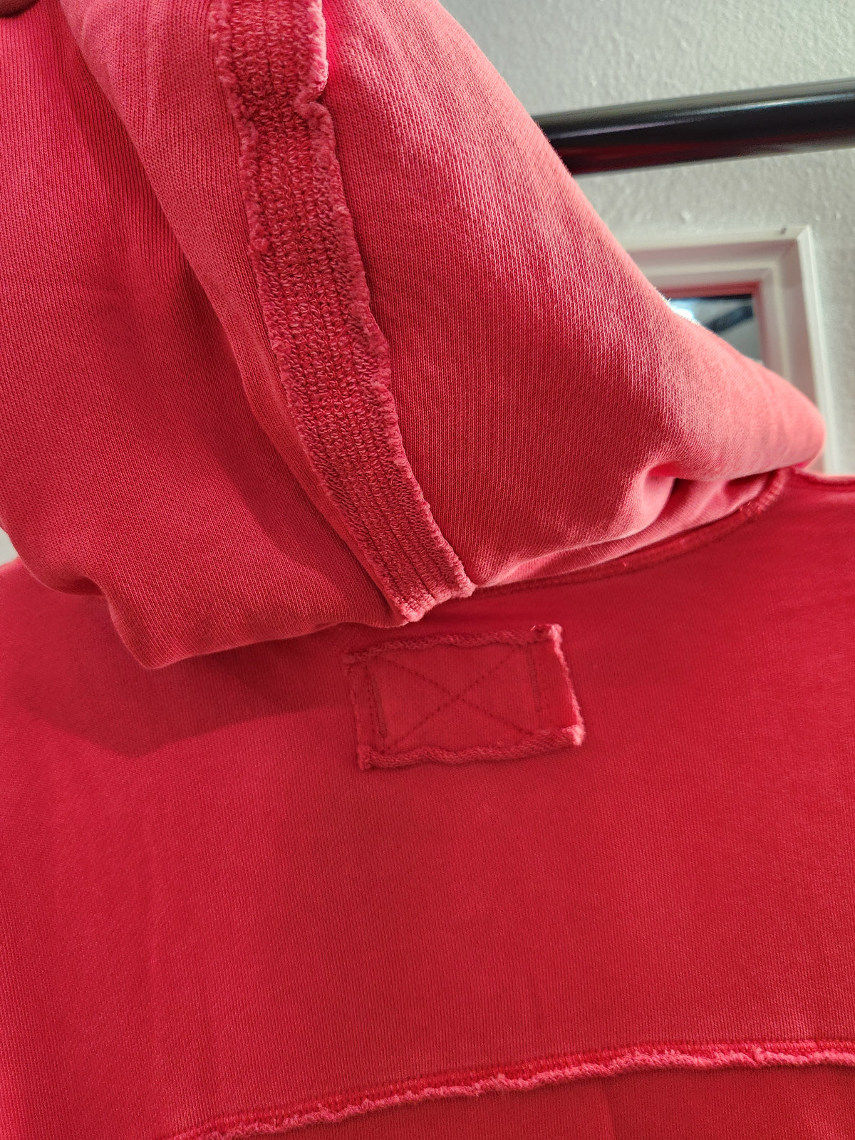 A close-up of a red hooded sweatshirt showing the hood lining, ribbed collar, and a small rectangular embroidered patch on the chest.