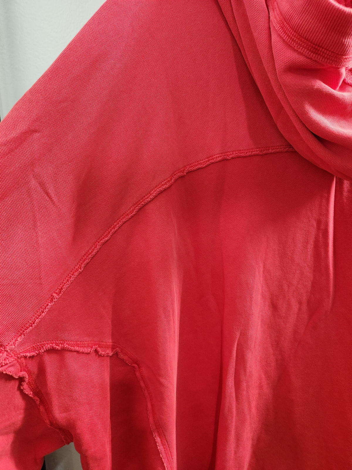 A close-up of a coral pink or red hooded sweatshirt, showing the shoulder seam, raw hem detail, and soft draped fabric folds.