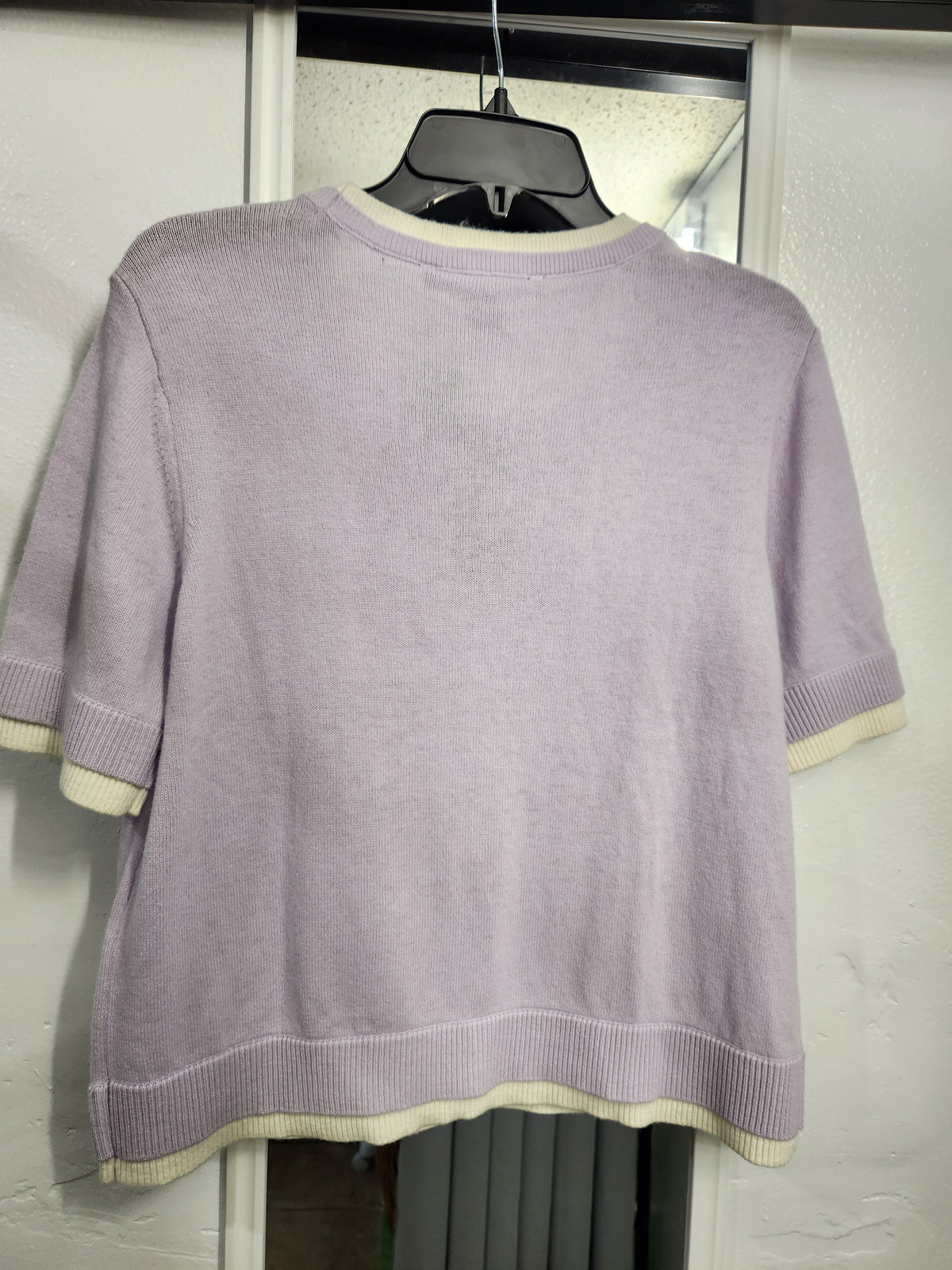 A light purple short-sleeve knit sweater with cream ribbed trim, hanging on a black hanger against a white door.