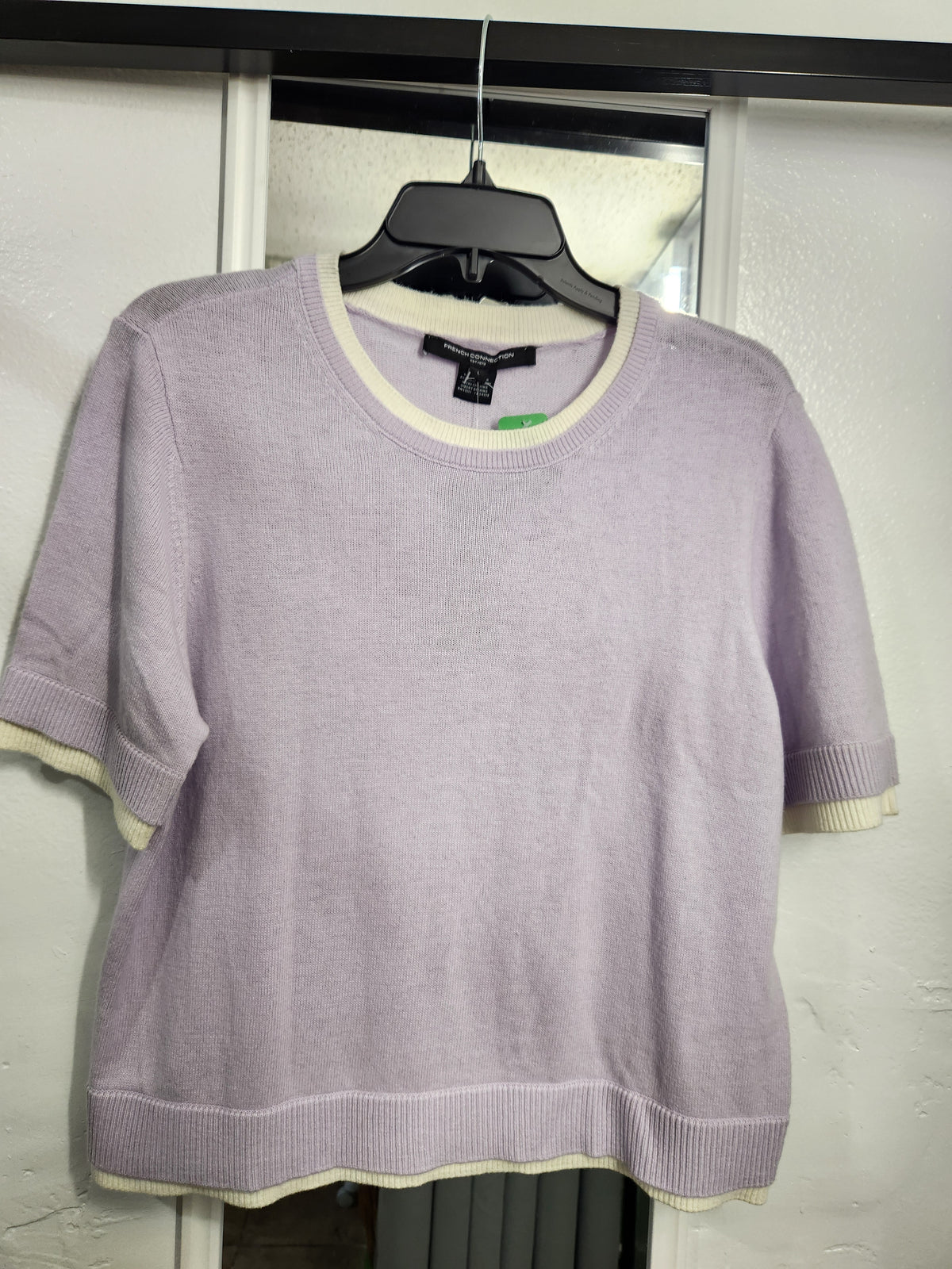 A light purple short-sleeve knit sweater with cream ribbed trim on the collar, sleeve cuffs, and hem, hanging on a black hanger against a white door.