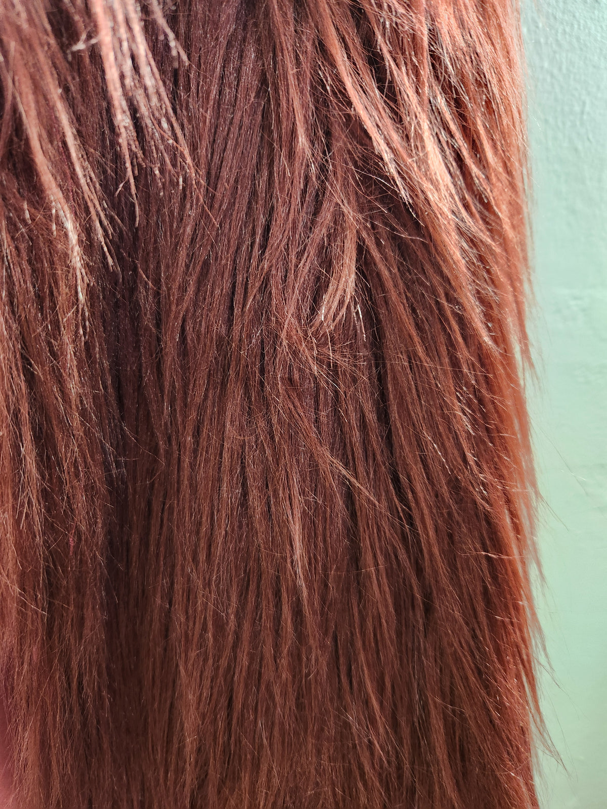 A close-up of straight, shoulder-length hair in a warm coppery auburn shade, with lighter golden highlights, set against a pale mint green background.