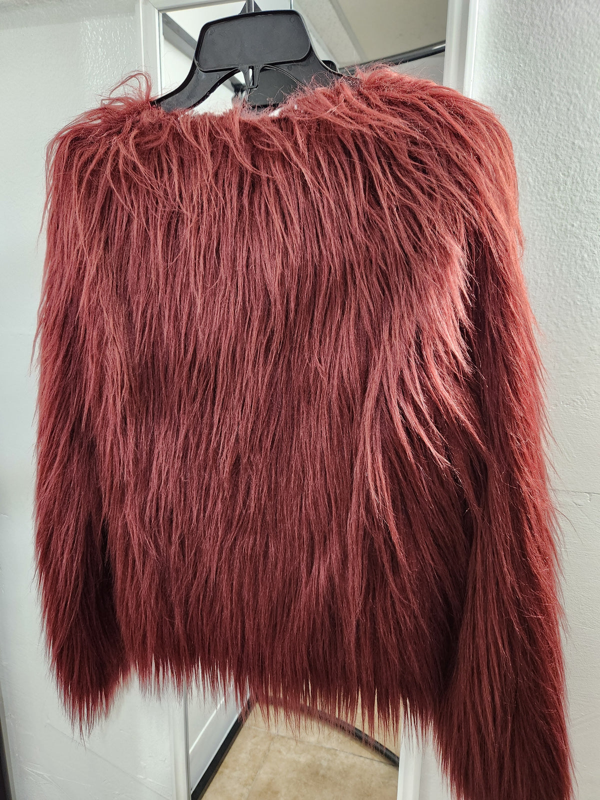 A shaggy, long-pile faux fur sweater in a dusty rose or muted burgundy color, hanging on a black hanger against a white door frame.