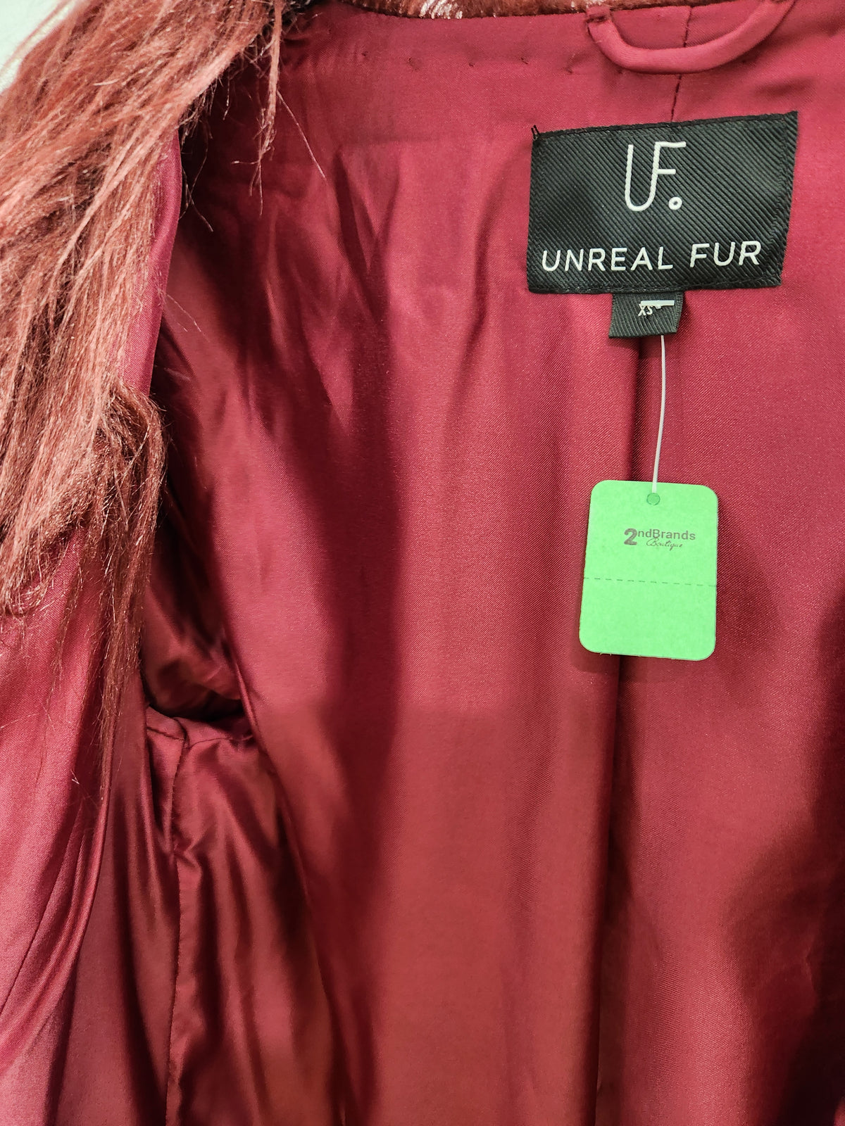 Close-up of a burgundy satin-lined jacket or coat interior with a black 'UF. UNREAL FUR' brand label and a bright green hangtag.