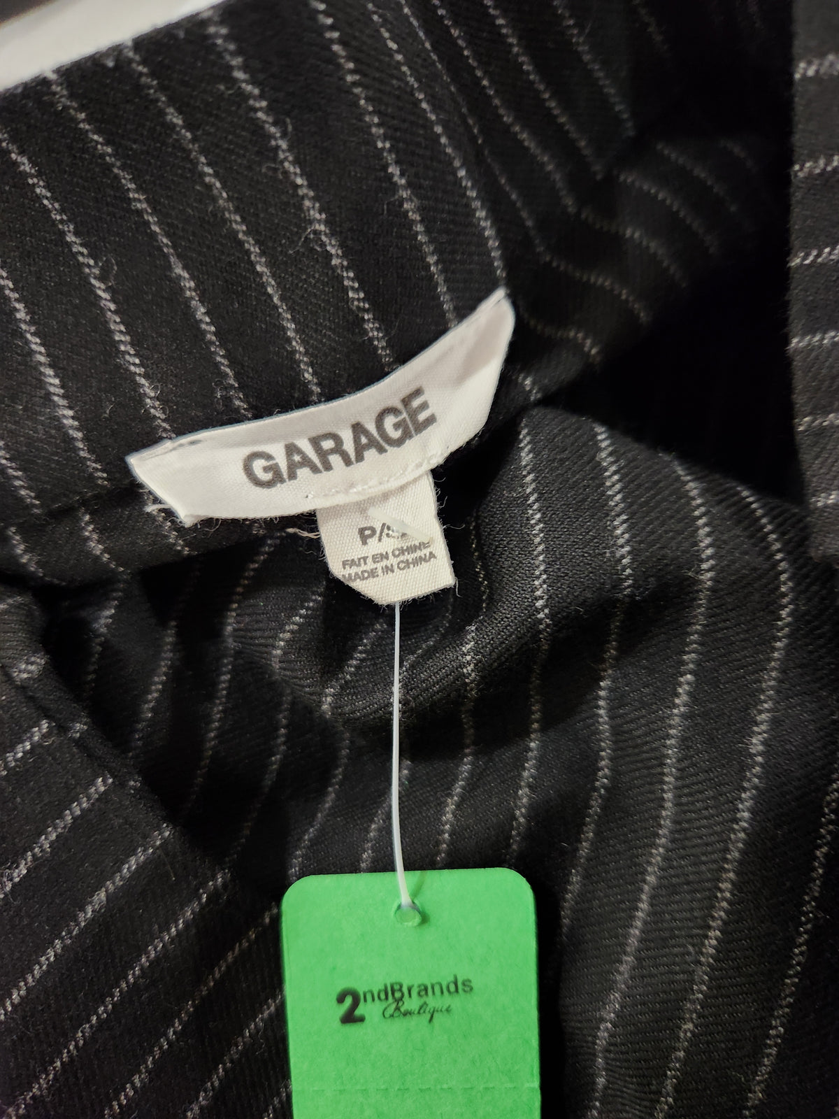 Close-up of a black pinstriped garment with a visible white interior neck label reading GARAGE and a green retail hangtag below it.