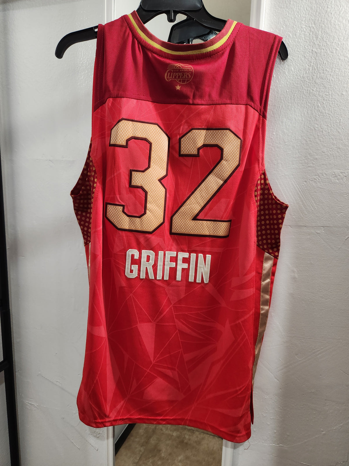 A red sleeveless basketball jersey hanging on a black hanger against a white wall. The back of the jersey features the number 32 in beige with black outlines, the name GRIFFIN in white, and a subtle geometric pattern.