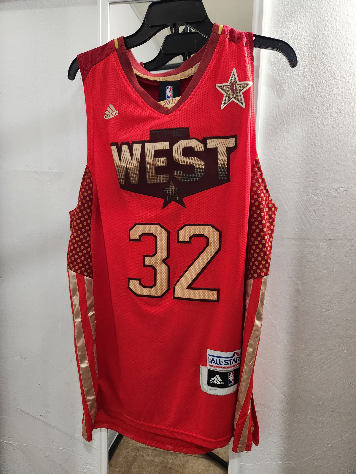 A red Adidas All-Star basketball jersey hanging on a black hanger against a white wall. The jersey features 'WEST' and the number '32' in gold and black, with a gold star logo on the upper right chest and an All-Star patch on the lower right hem.