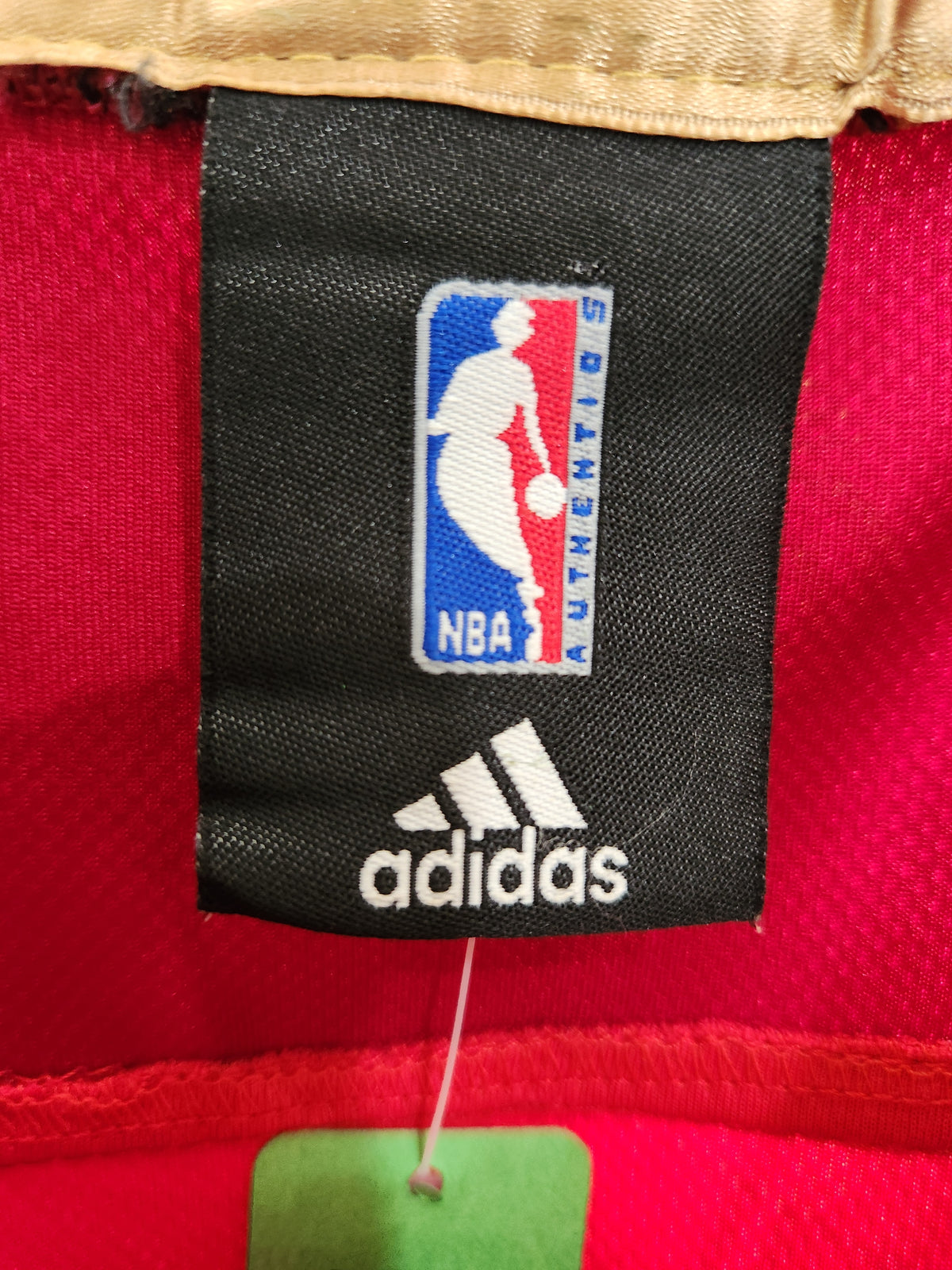 Close-up of an Adidas NBA Authenticities label sewn onto a red athletic jersey, showing the black woven tag with white Adidas branding and the blue/red/white NBA logo.