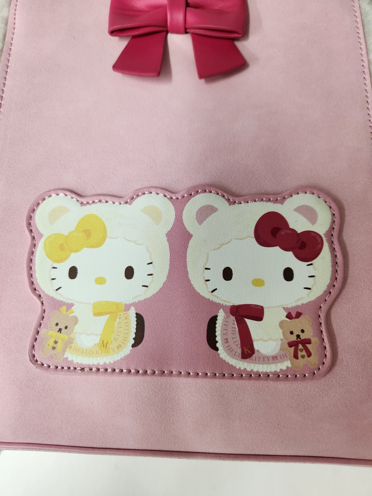 A pink faux-leather bag featuring two Hello Kitty character patches on the front, one with a yellow bow and dress and one with a red bow and dress, plus a matching pink bow at the top.