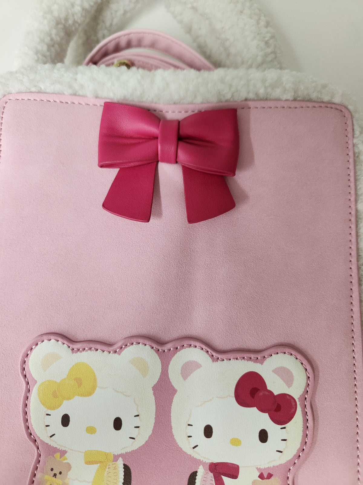 A pink faux-leather backpack featuring a large pink bow on the upper front and two Hello Kitty character patches at the bottom, with a white sherpa lining visible along the top edge.