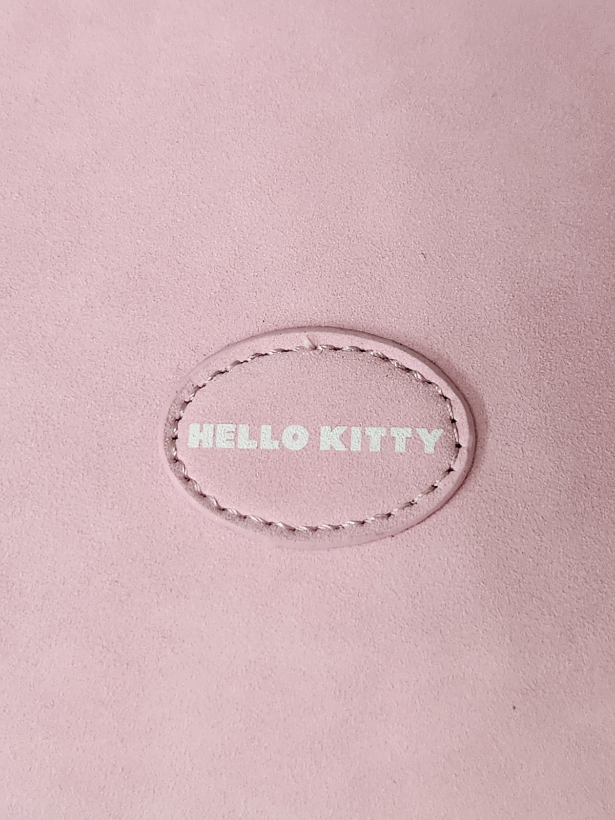 A close-up of a dusty pink leather surface featuring an oval stitched patch with the text 'HELLO KITTY' in a light pink tone.