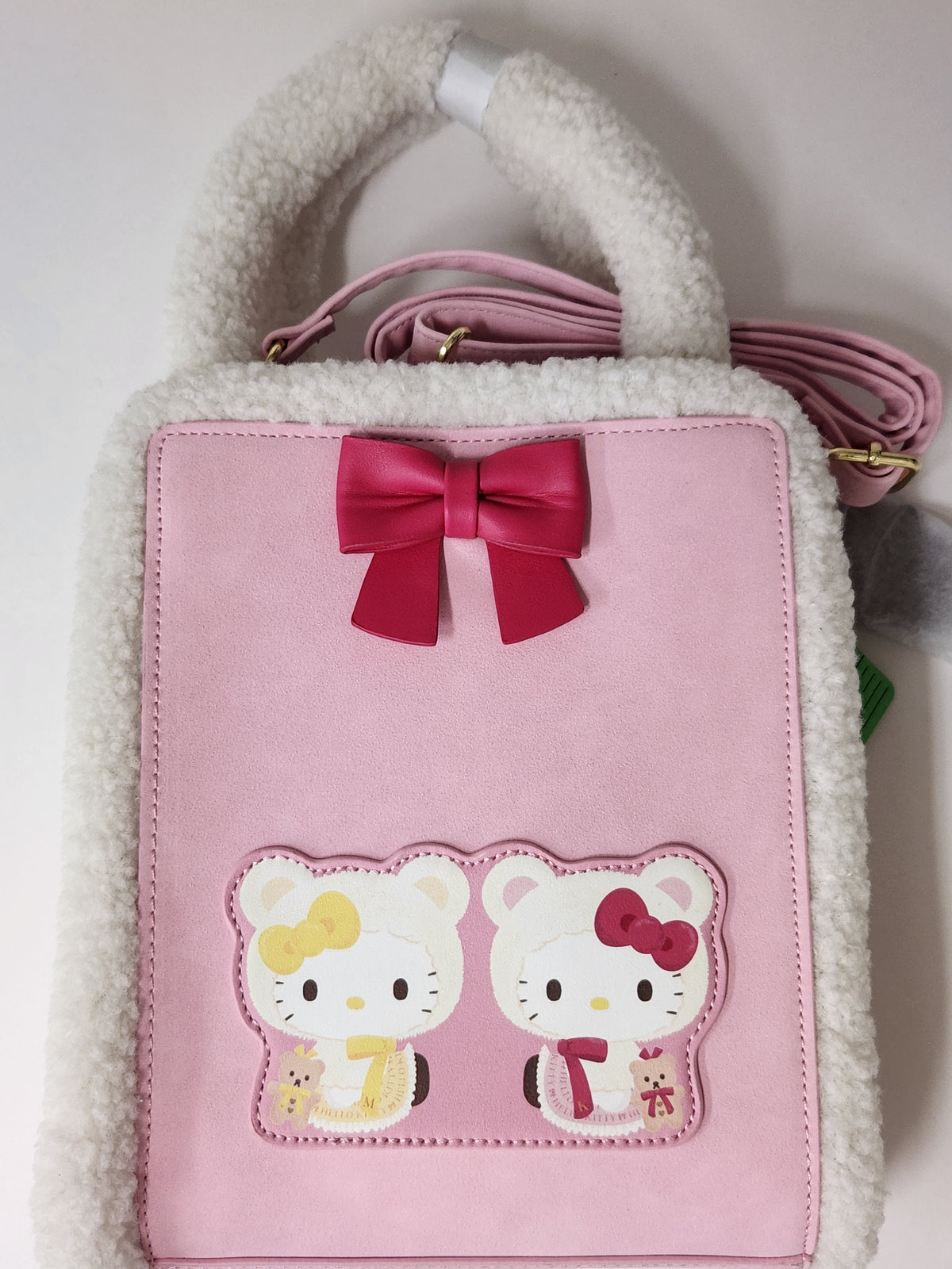 A pink faux-leather handbag with a white sherpa fleece handle and trim, featuring a dark pink bow on the front and two Hello Kitty patches. A pink shoulder strap is visible behind the bag.