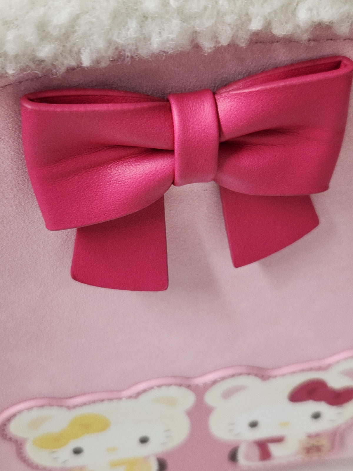 A close-up of a pink faux-leather bow accessory attached to a light pink fabric item with a white faux-fur trim and Hello Kitty character graphics visible at the bottom.