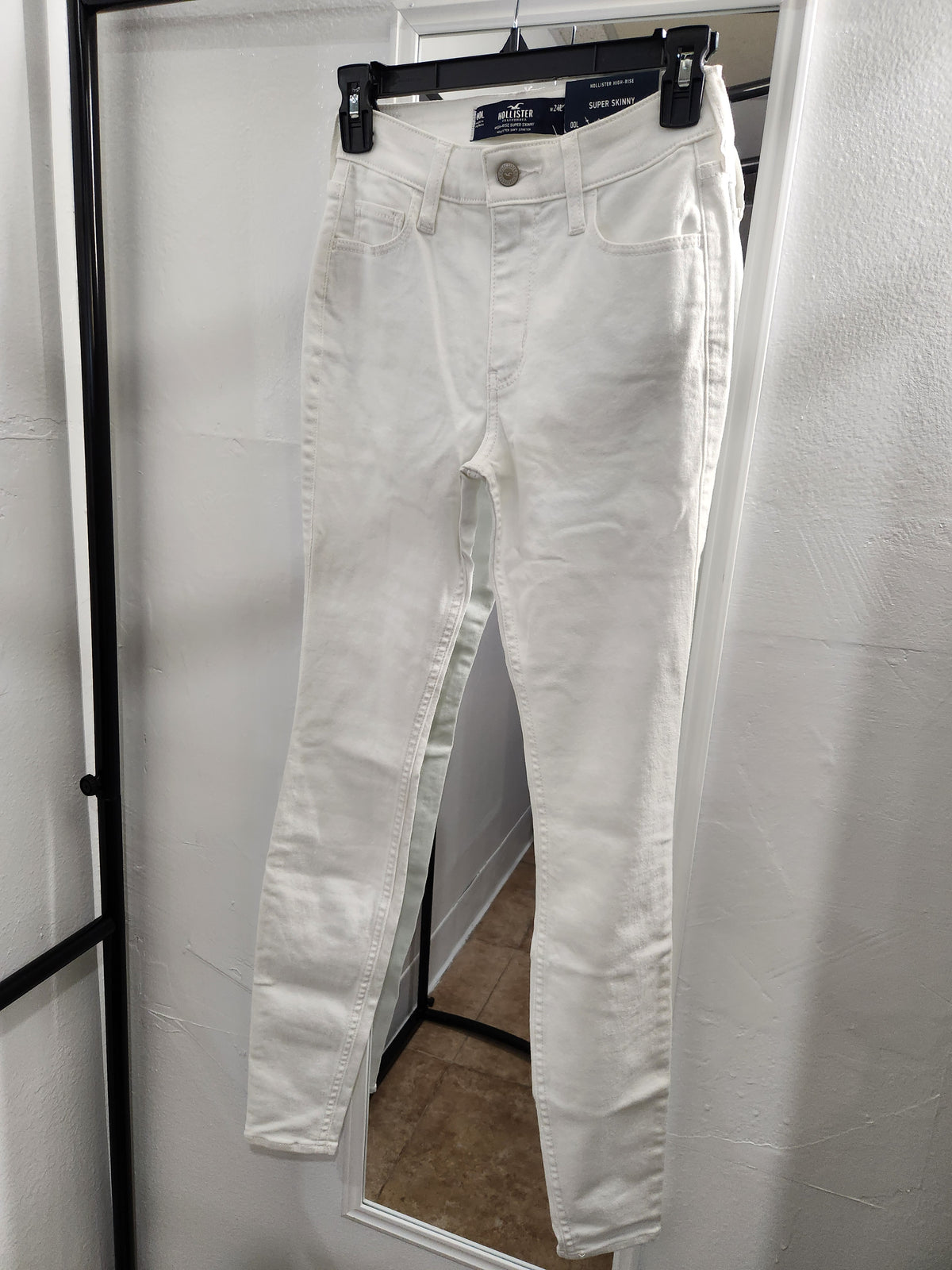 White slim-fit denim jeans hanging on a black hanger against a white wall, with a mirror reflecting a home interior background.