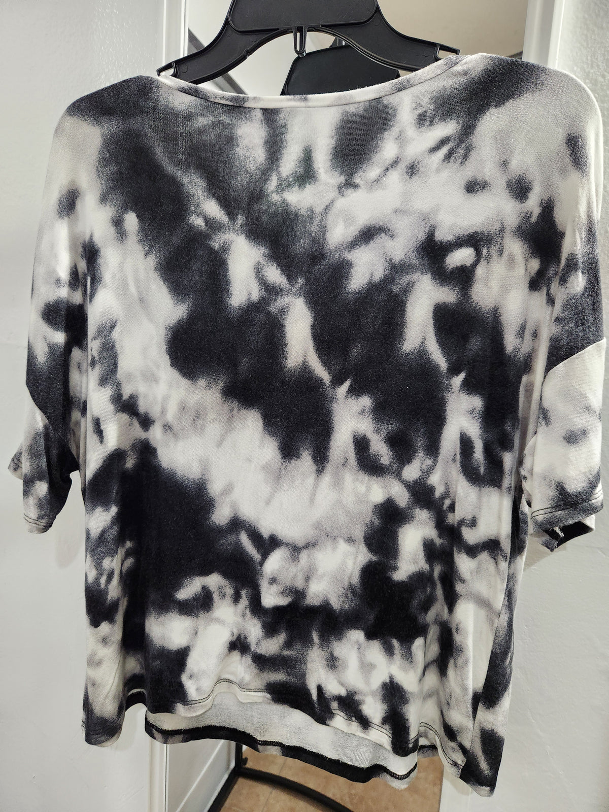 A black and white tie-dye short-sleeve top hanging on a black hanger against a white door background.