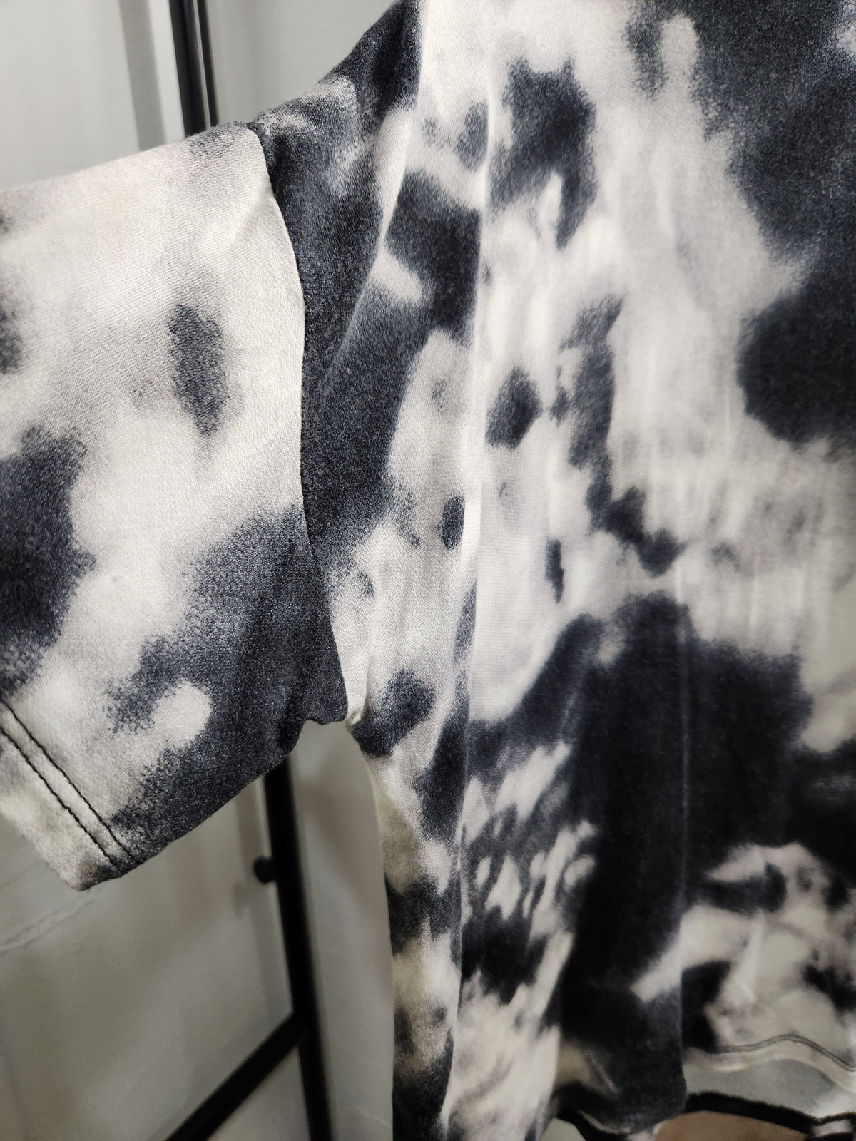 A close-up of a short-sleeve tie-dye t-shirt featuring a black and white mineral-wash or bleach-dyed pattern, shown hanging on a black clothing rack.