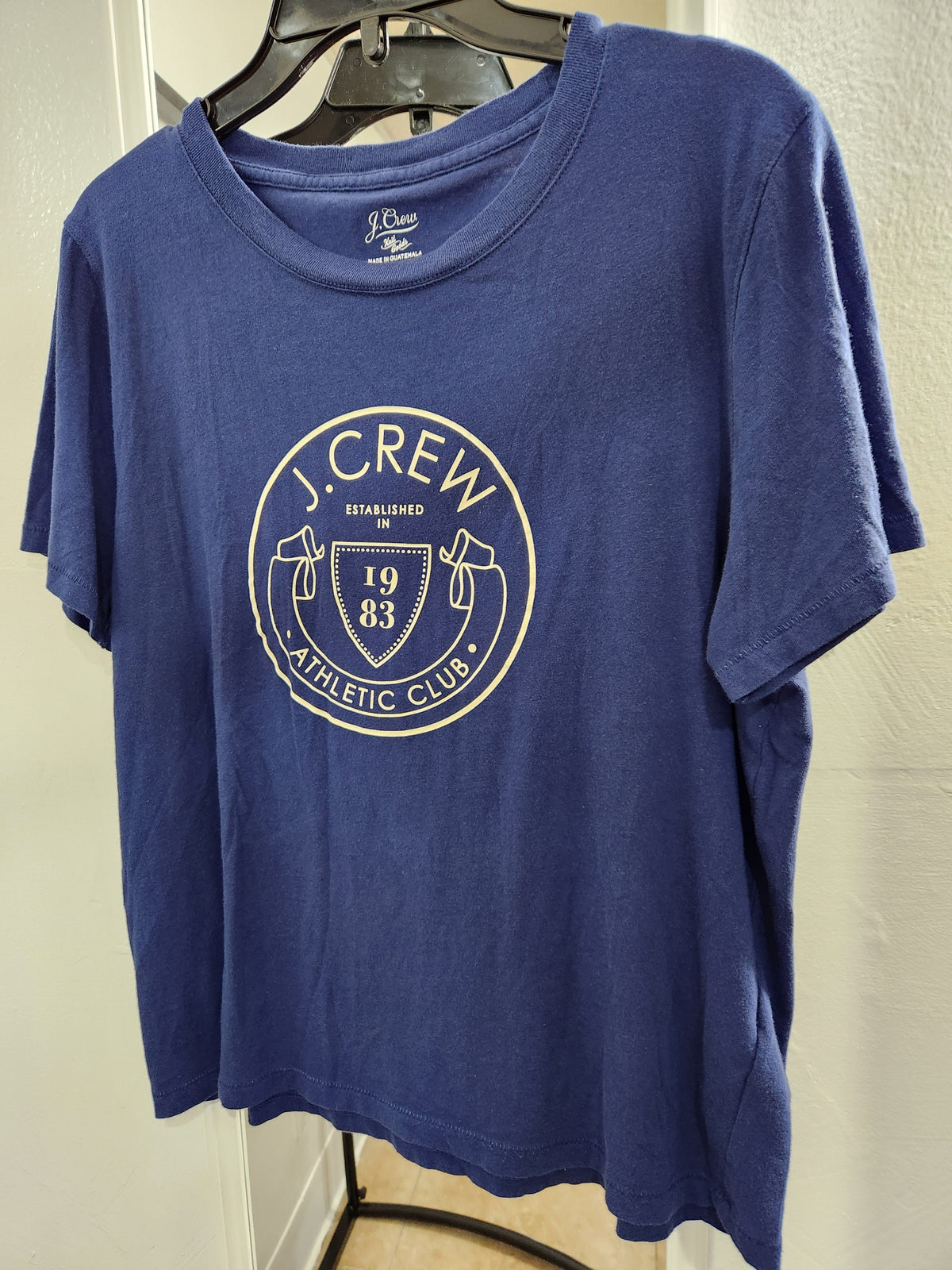 A blue short-sleeve crewneck t-shirt hanging on a black hanger against a light-colored wall. The shirt features a circular white graphic on the chest with the text 'J.