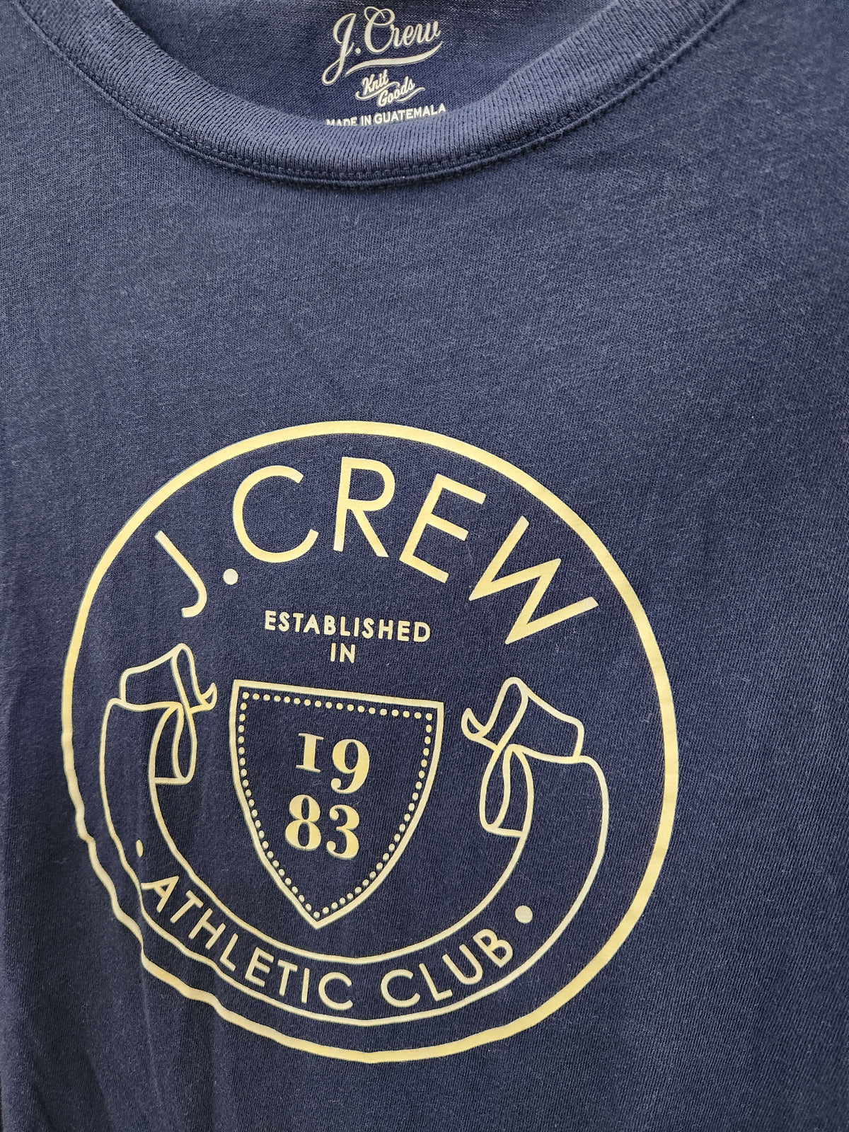Close-up of a navy blue heathered t-shirt featuring a circular gold graphic print on the front with the text 'J. CREW ESTABLISHED IN 1983 ATHLETIC CLUB'.