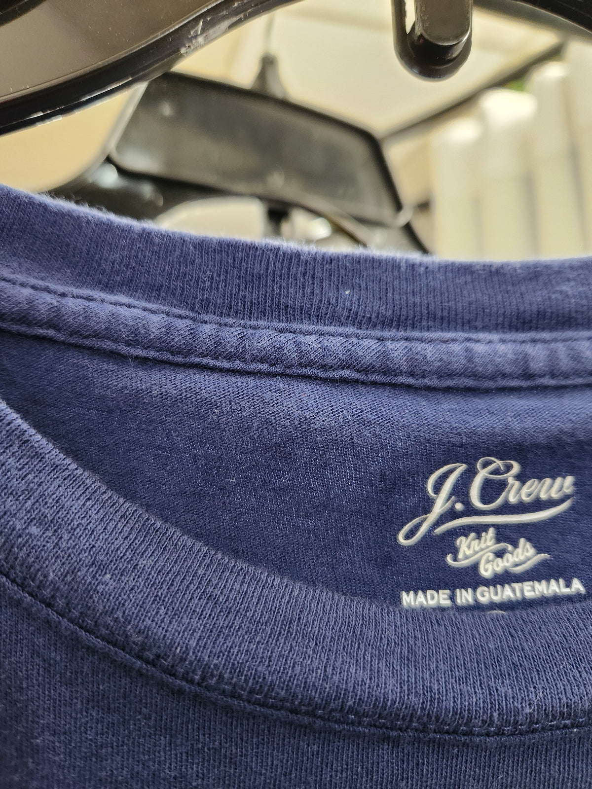 Close-up of a navy blue crewneck t-shirt hanging on a black hanger, showing the white embroidered J.Crew brand logo and 'Made in Guatemala' text inside the collar.
