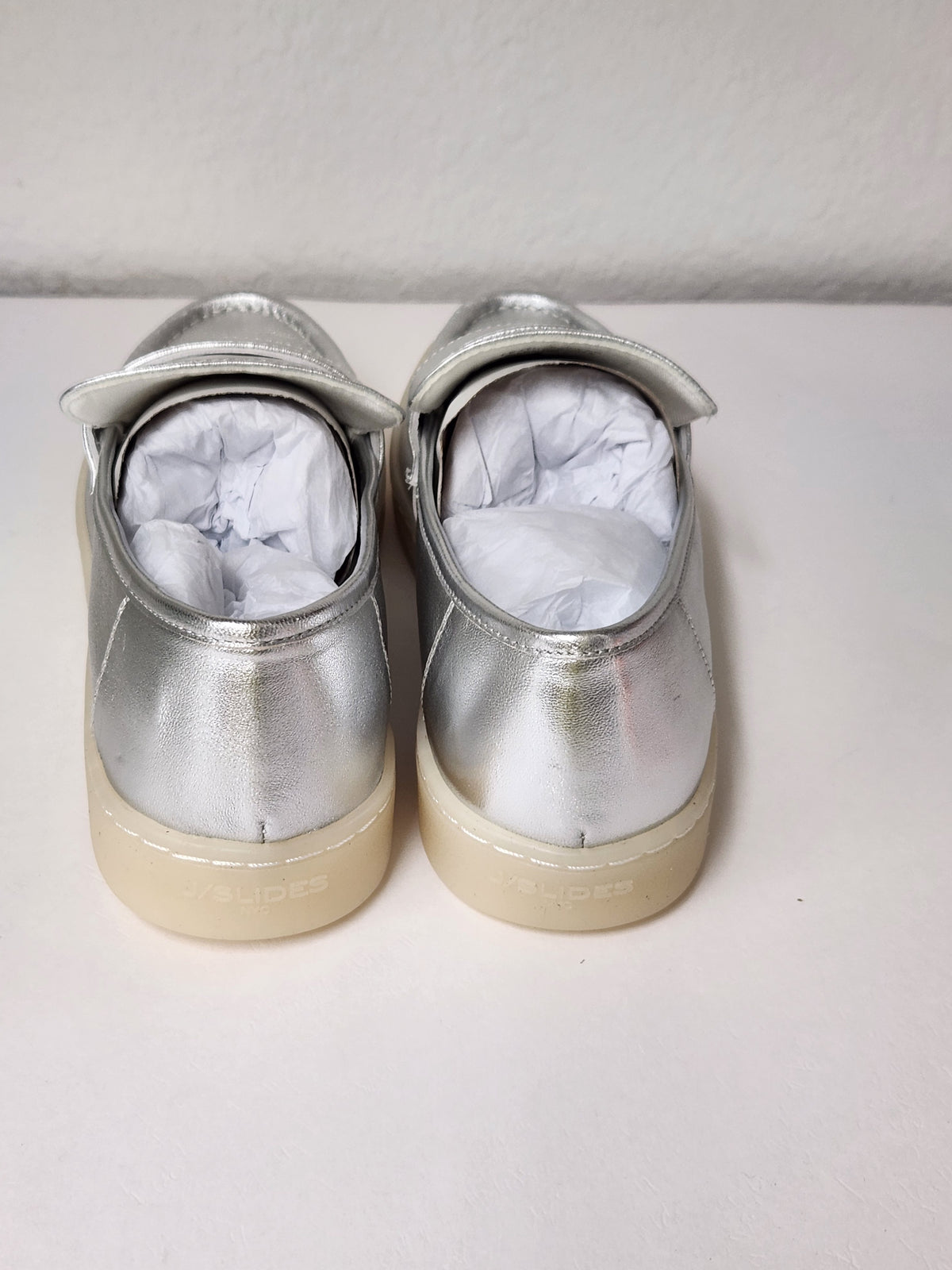 A pair of metallic silver Mary Jane shoes with beige soles, shown from the back with white paper stuffing inside, on a light surface against a white wall.