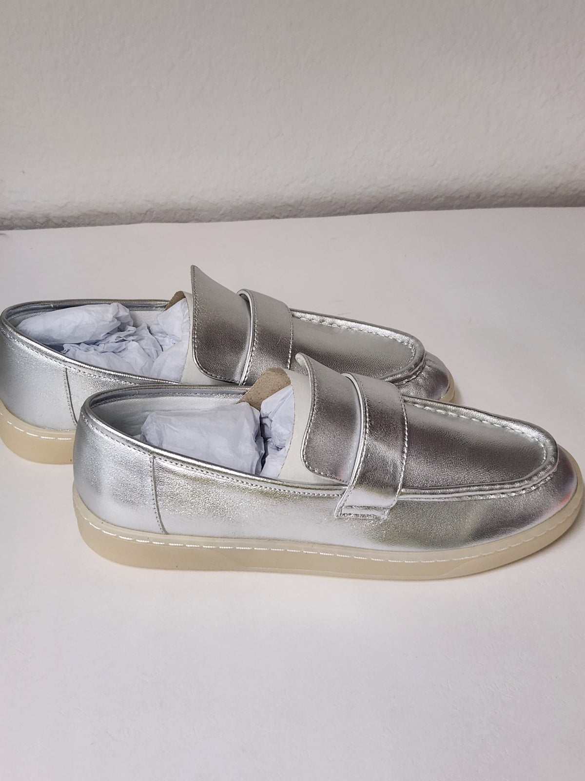 A pair of metallic silver slip-on loafers with a single wide strap across the top and a light beige sole, shown on a plain white surface.