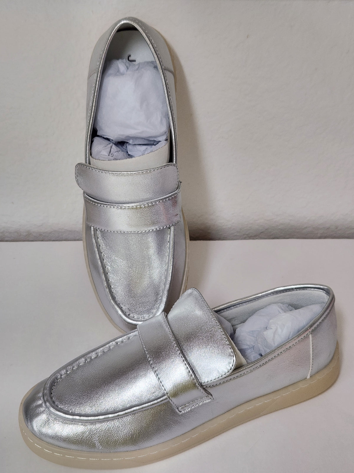 A pair of metallic silver slip-on loafers with a single strap across the instep and a beige sole, shown on a plain light background.