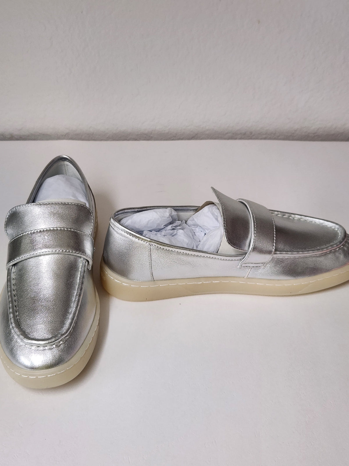 A pair of metallic silver slip-on loafers with cream-colored soles, shown on a plain off-white surface against a light gray wall.