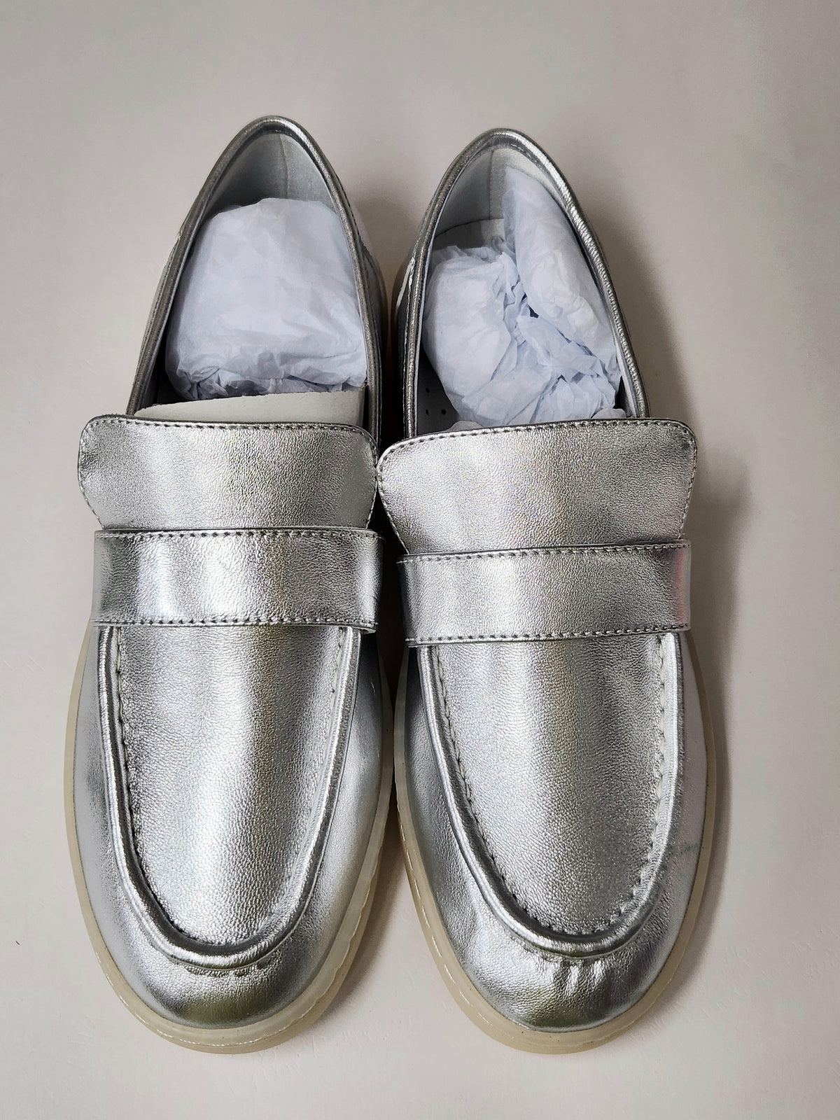 A pair of metallic silver slip-on loafers with a wide strap across the instep, photographed from above on a light beige background. White tissue paper is visible inside the shoes.