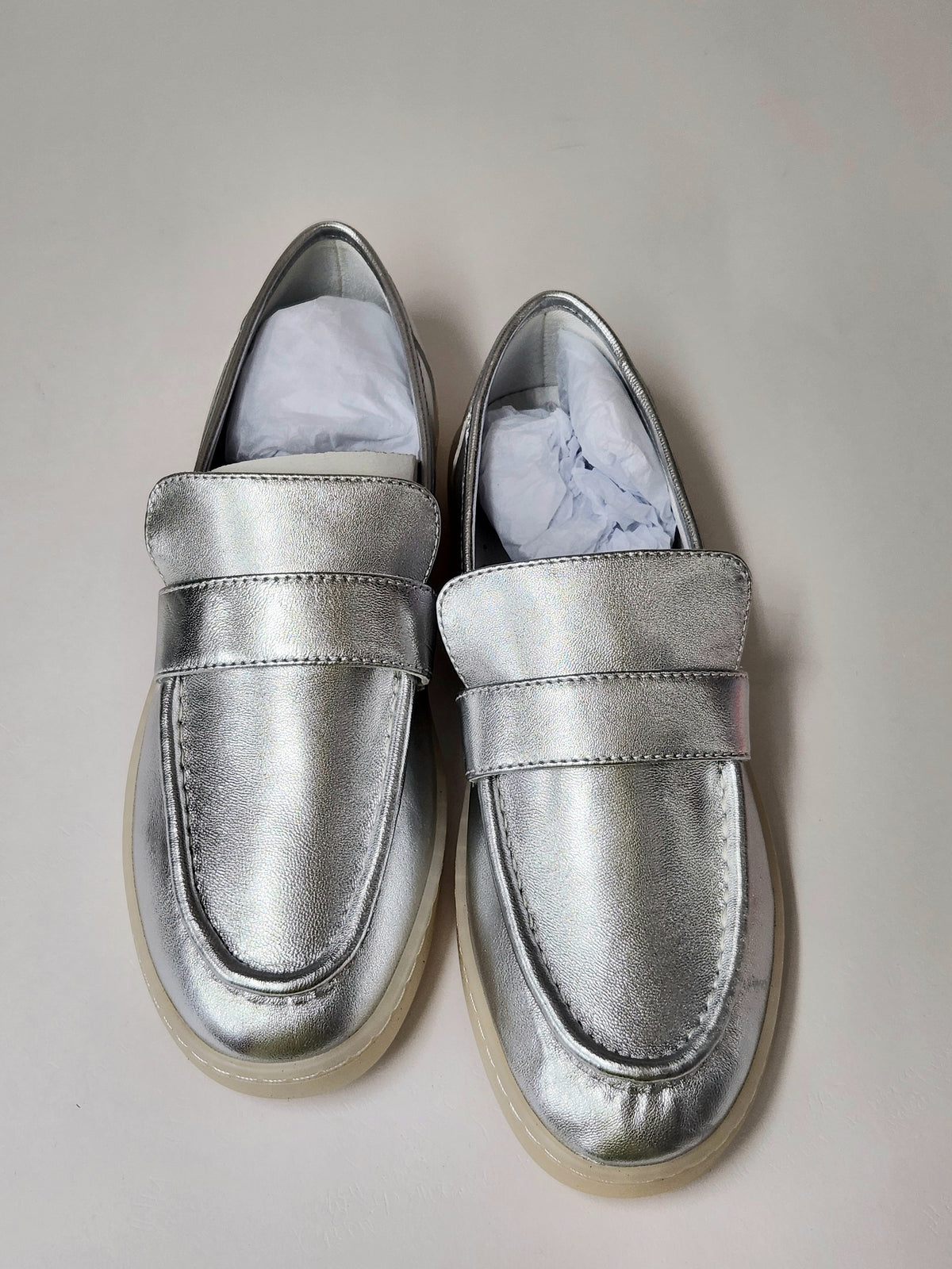 A pair of metallic silver slip-on loafers with a wide strap and a thick beige sole, shown on a light gray background with white tissue paper inside.
