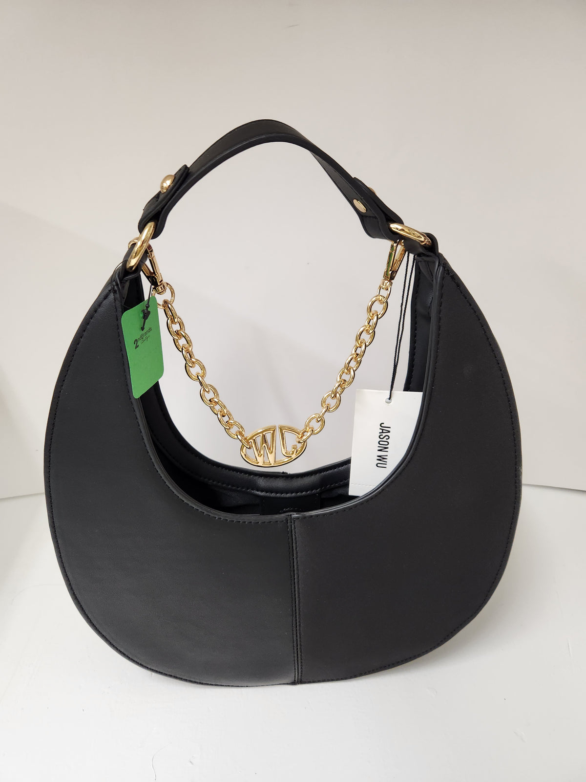 A black leather crescent-shaped shoulder bag with gold-tone hardware, featuring a gold chain detail with a central logo clasp, a top handle, and retail tags attached, set against a plain white background.