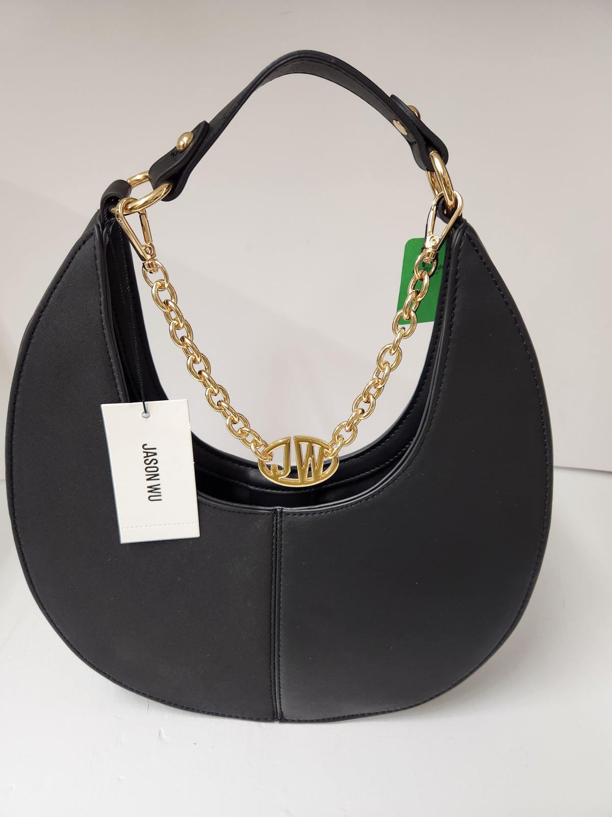 A black leather crescent-shaped shoulder bag with a gold chain link detail and a gold monogrammed O-shaped charm. It features a black leather top handle, a white paper tag reading 'JASON NU', and a small green hangtag, photographed on a light gray background.