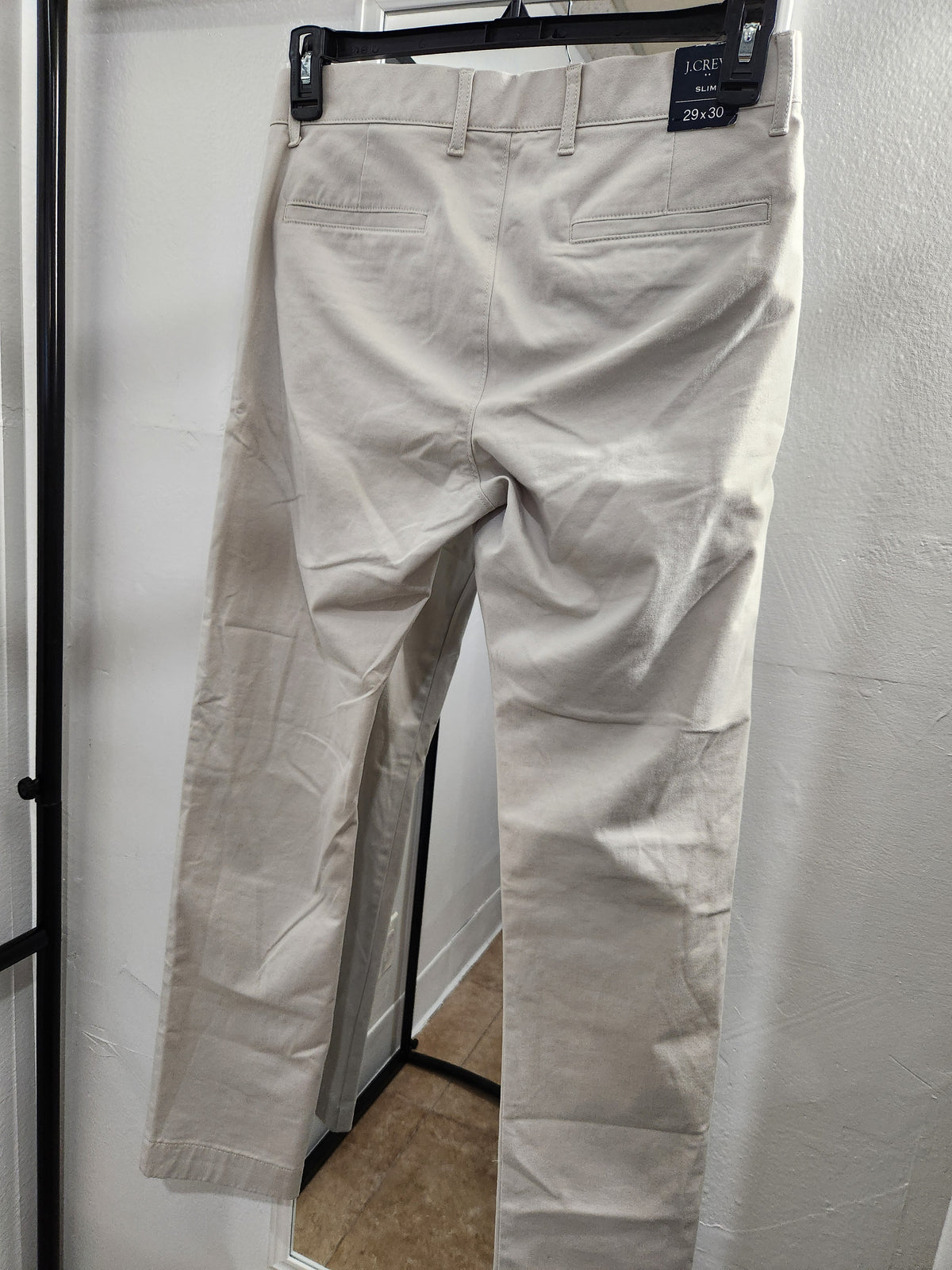 A pair of light beige chino pants hanging on a black hanger against a white wall, showing the back pockets and waistband details.