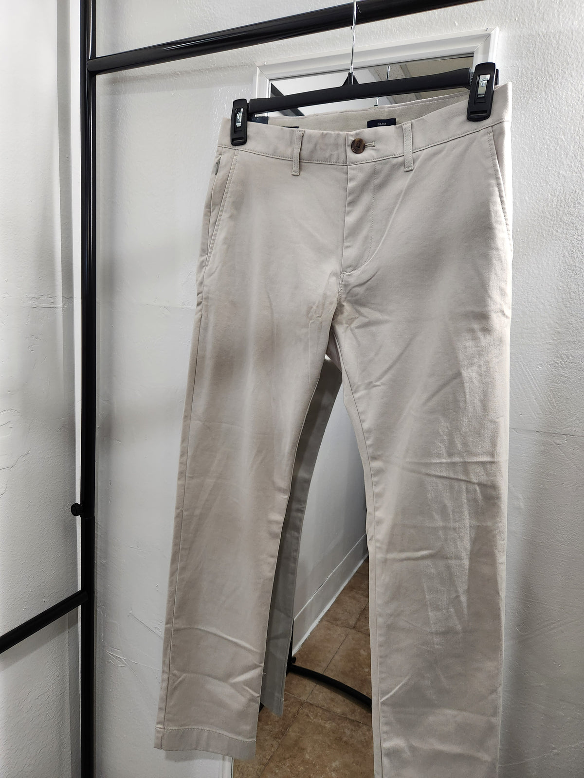 A pair of light beige chino pants hanging on a black hanger from a clothing rack, set against a white wall.