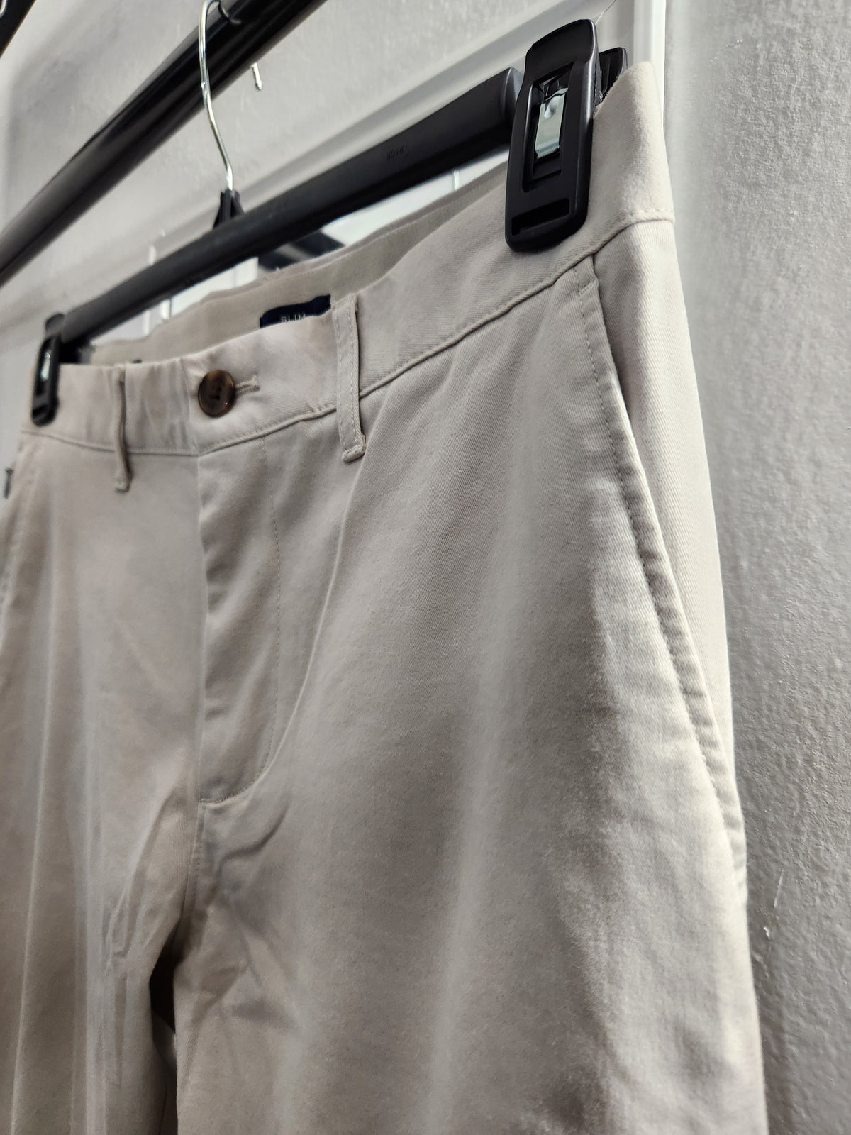 Beige chino pants hanging on a black hanger against a light gray wall.