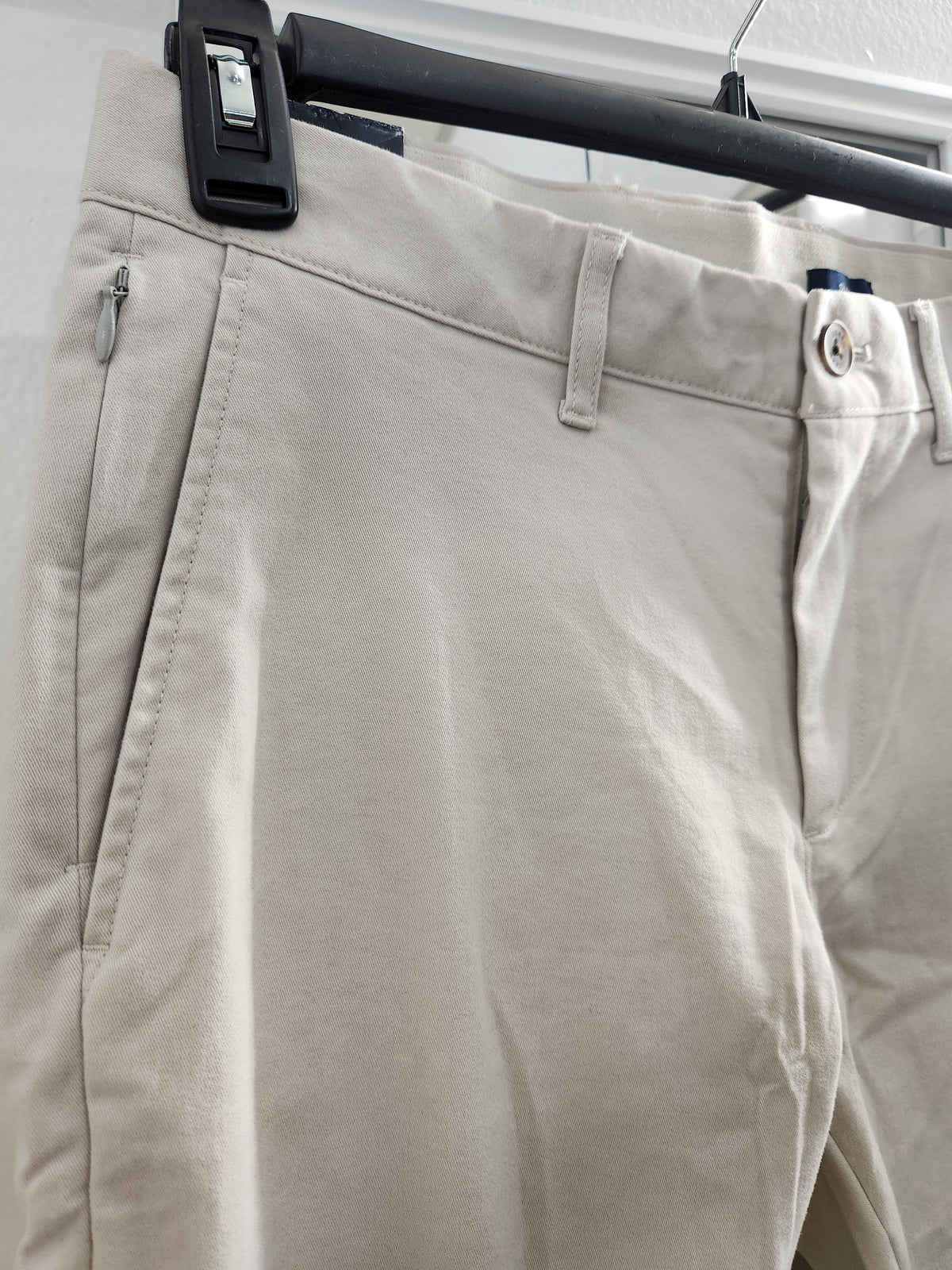Beige chino pants hanging on a black hanger, showing the front waistband, belt loops, button, and side pocket details.