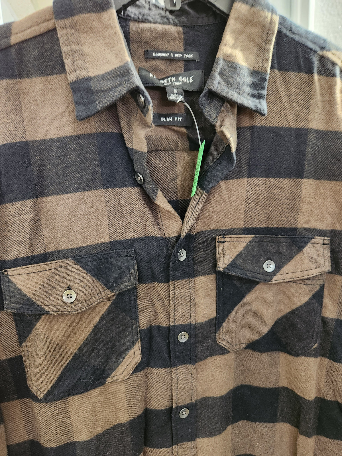 A close-up of a North Sole plaid button-down shirt in brown and dark navy blue, featuring two chest pockets and a green price tag.