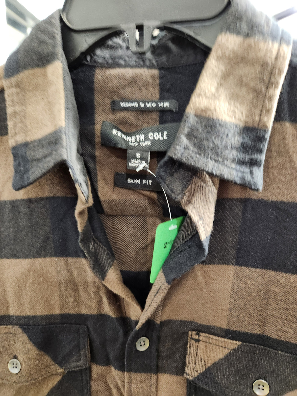 A close-up of a Kenneth Cole Slim Fit flannel shirt in a brown, beige, and black plaid pattern, hanging on a black hanger with a green size tag.