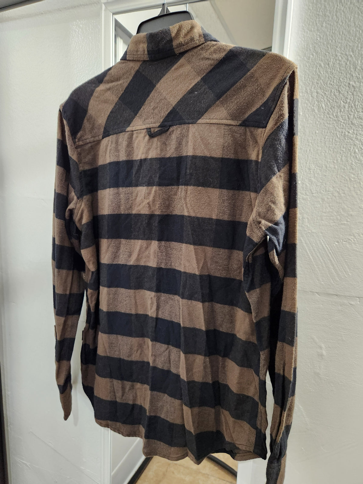 A long-sleeve plaid button-up shirt in brown and black, hanging on a black hanger against a white door and textured wall.