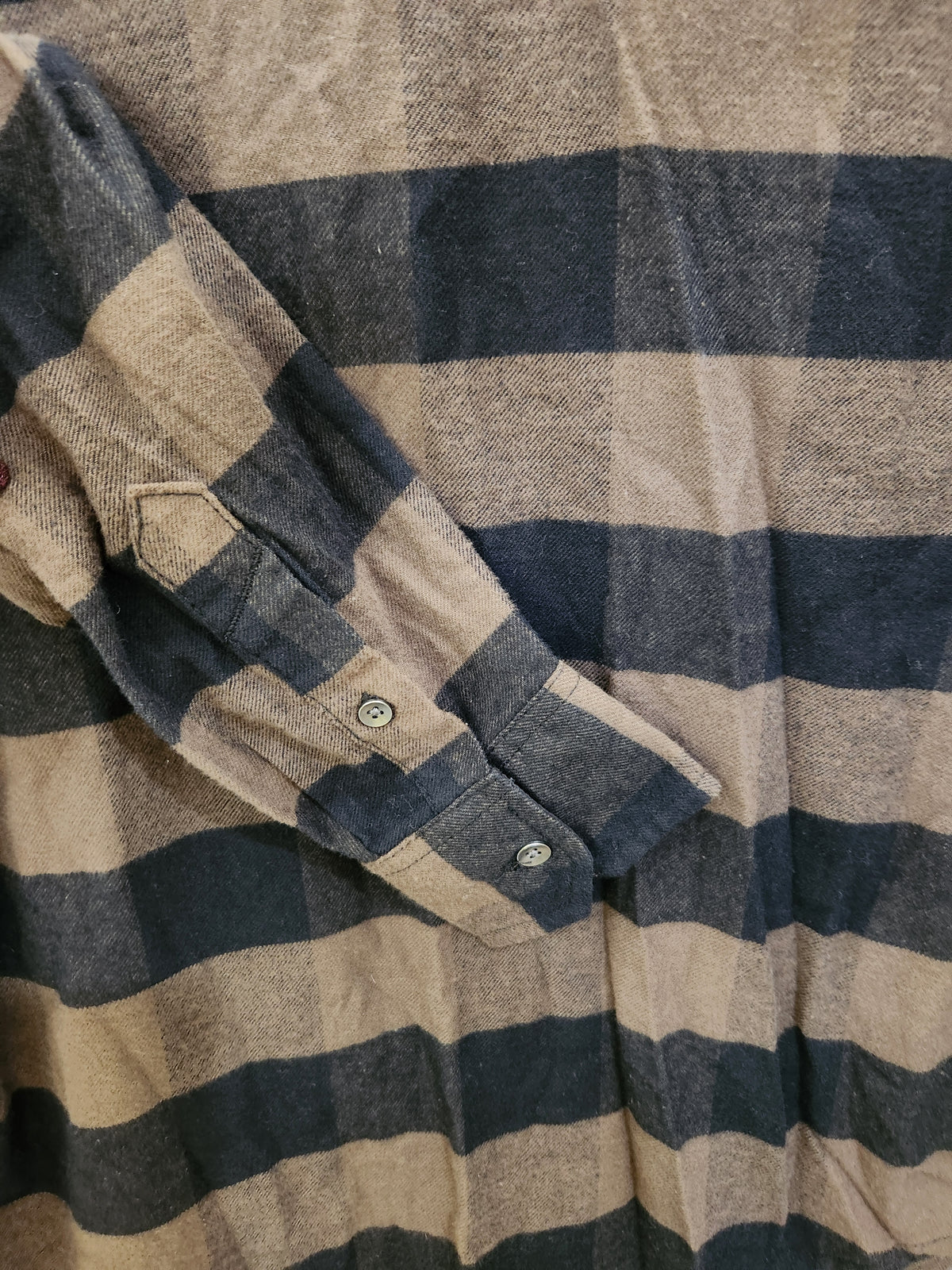 A close-up of a plaid flannel shirt with horizontal stripes in navy blue and a warm beige/tan color, showing the fabric texture and a buttoned pocket on the sleeve.