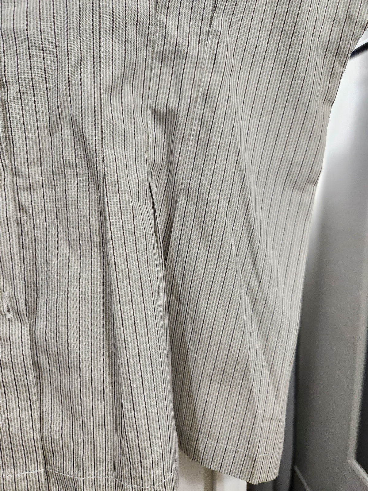 Close-up of a light beige or off-white fabric with thin vertical dark gray pinstripes, showing the texture and hem detail.