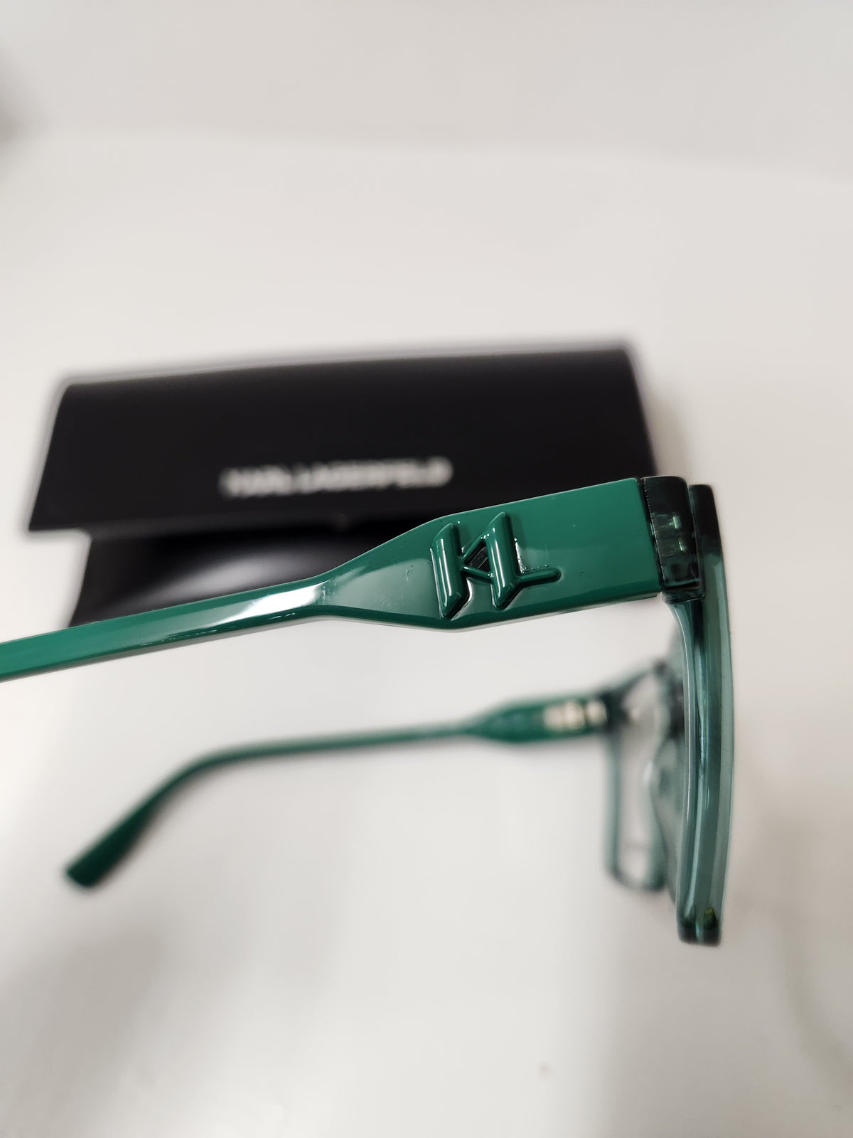 Green Karl Lagerfeld eyeglasses with a matching black hard case in the background, shown on a white surface.