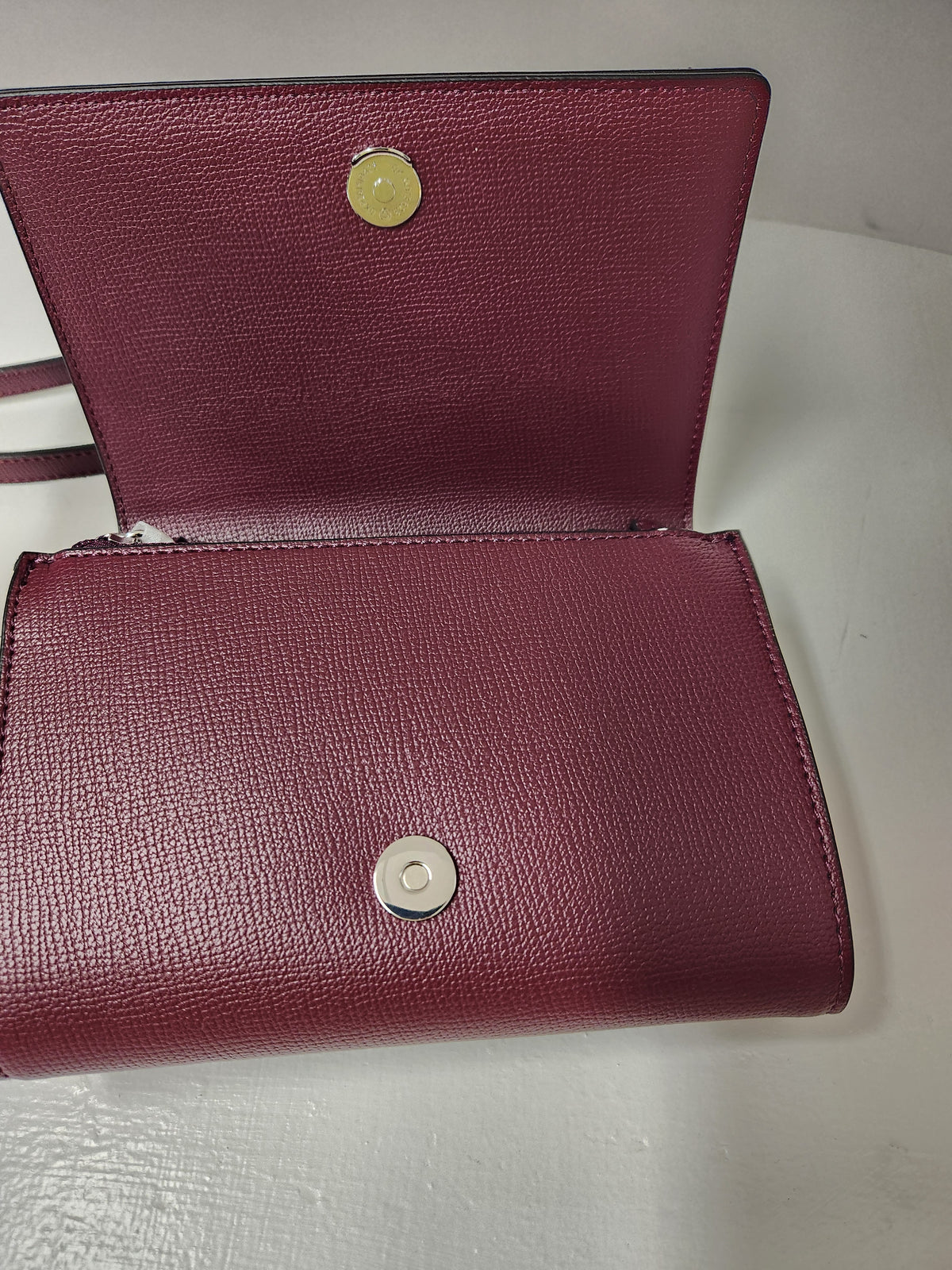 A burgundy textured leather wallet shown open, displaying its interior lining and two silver-tone snap button closures on a light gray surface.