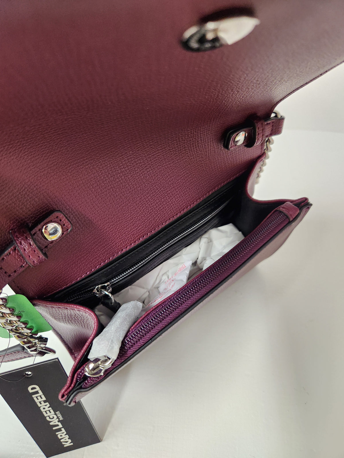 A close-up of a burgundy pebbled leather handbag shown open, revealing a black zippered interior compartment with white tissue paper inside. A black hangtag with the brand name 'HAUL LA SEED' is attached on the left side.