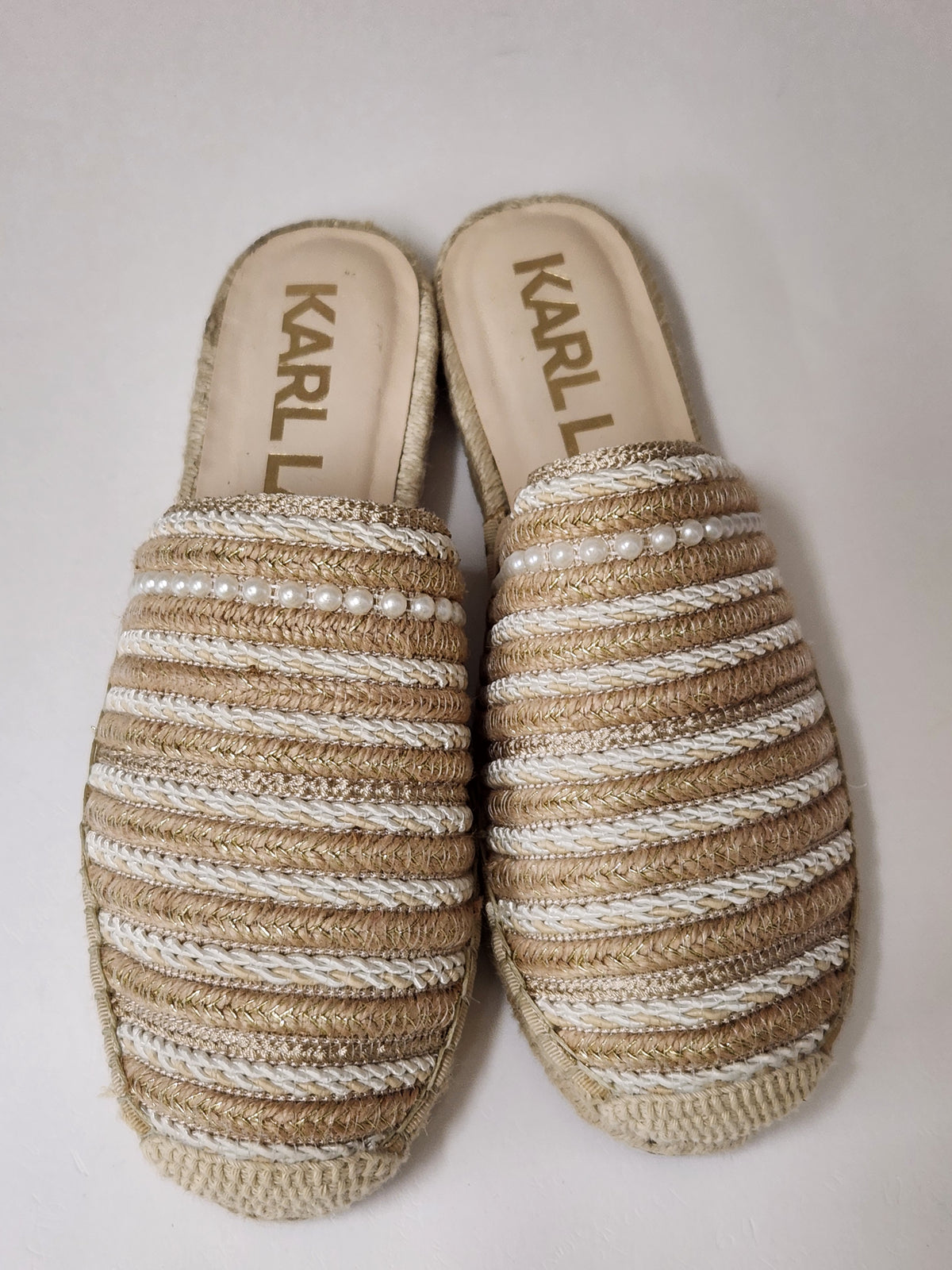 A pair of Kari L slip-on espadrille-style mule sandals with woven beige and off-white horizontal stripes and small pearl-like accents, shown on a plain light background.

