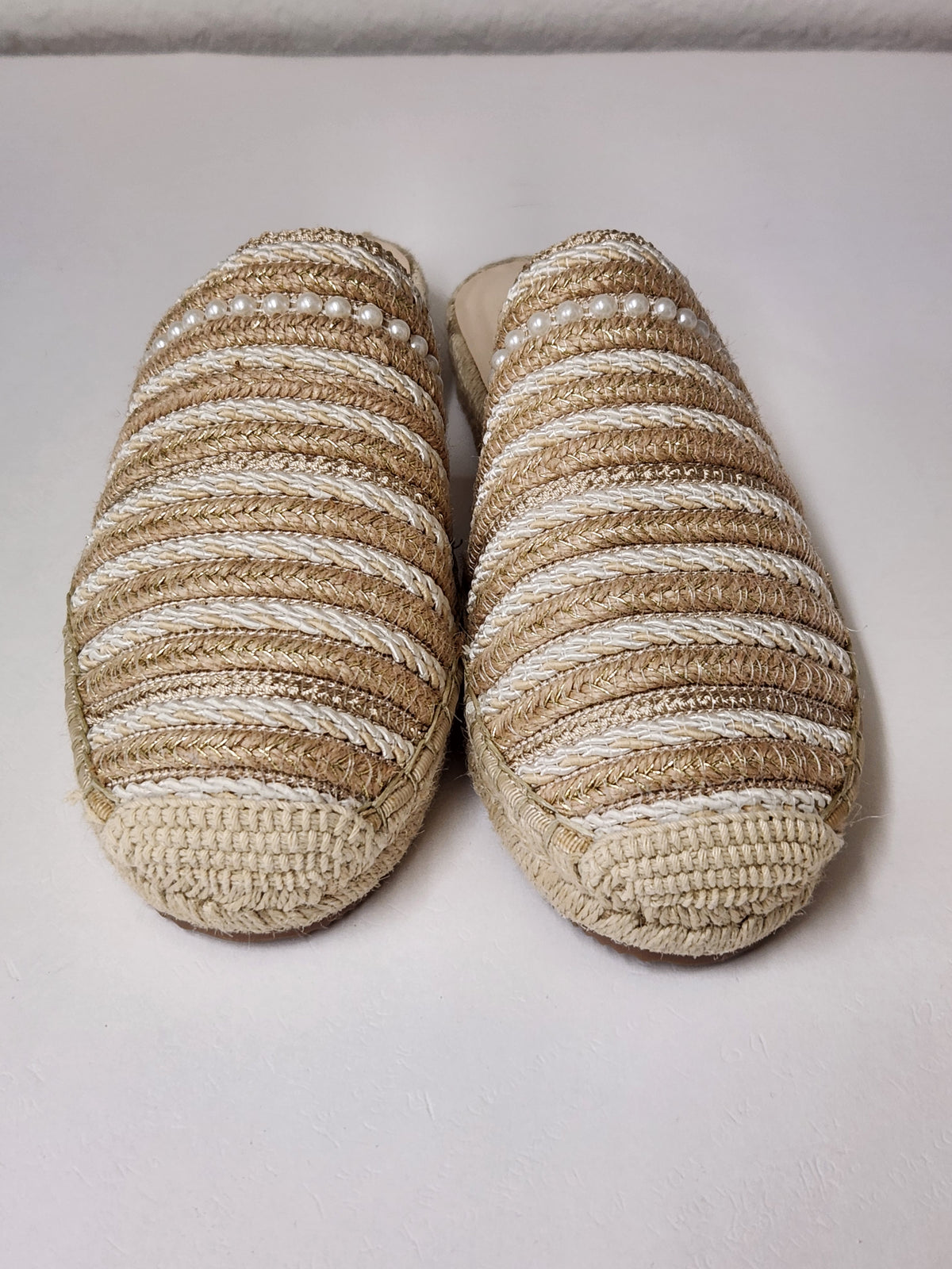 A pair of woven espadrille-style slip-on slippers with horizontal beige and off-white stripes, decorated with small white faux pearls across the upper, shown on a plain light background.