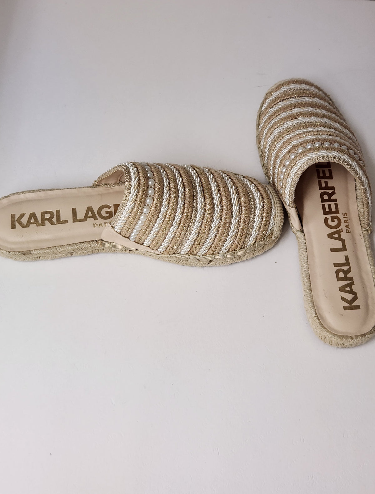 A pair of Karl Lagerfeld espadrille-style slide sandals with woven beige and white straps and a braided jute sole, displayed on a light off-white background.