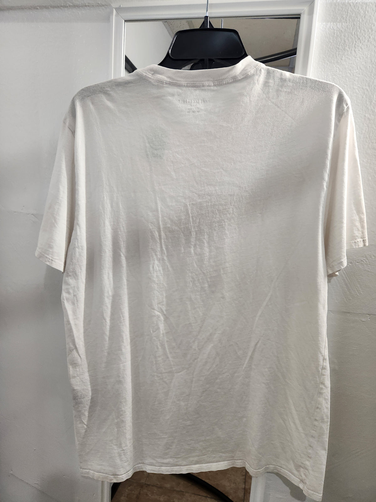 A white short-sleeve crewneck t-shirt hanging on a black hanger against a white wall.