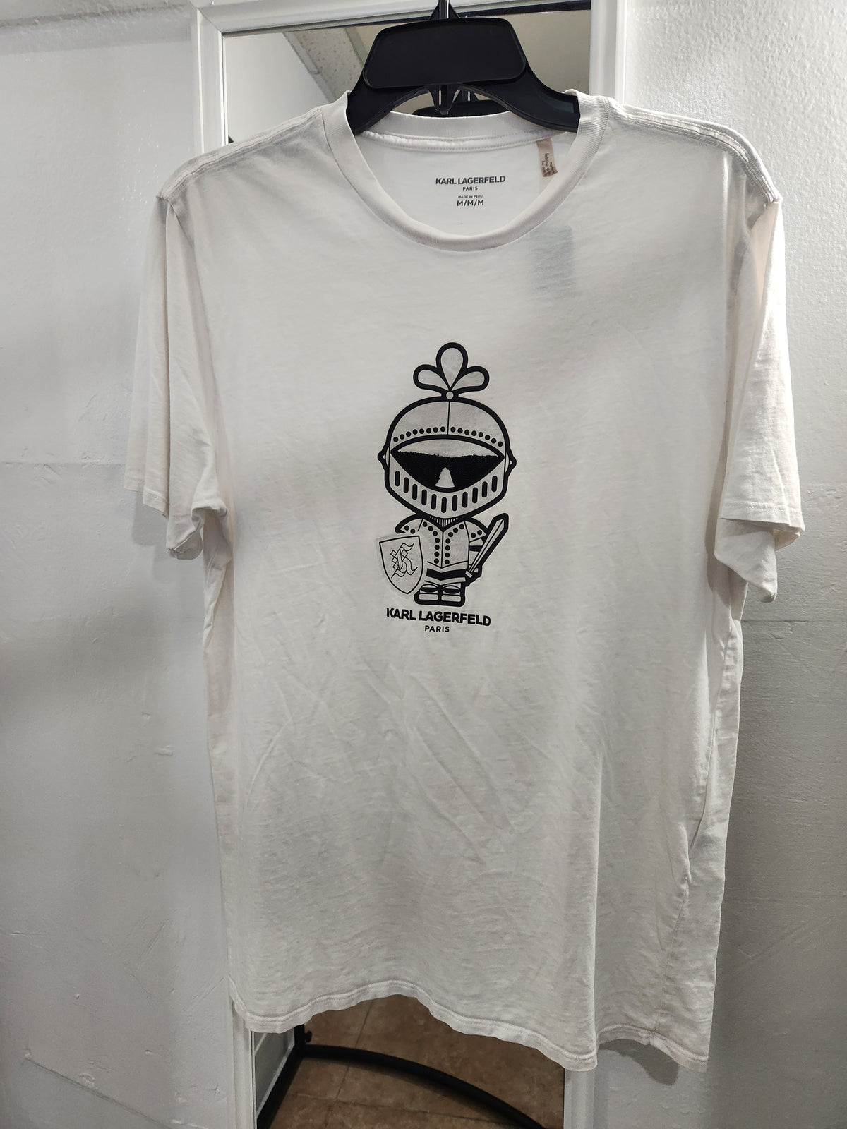 A white short-sleeve crewneck t-shirt by Karl Lagerfeld, featuring a black graphic of a knight in armor wearing a flower crown, hanging on a black hanger against a white wall.
