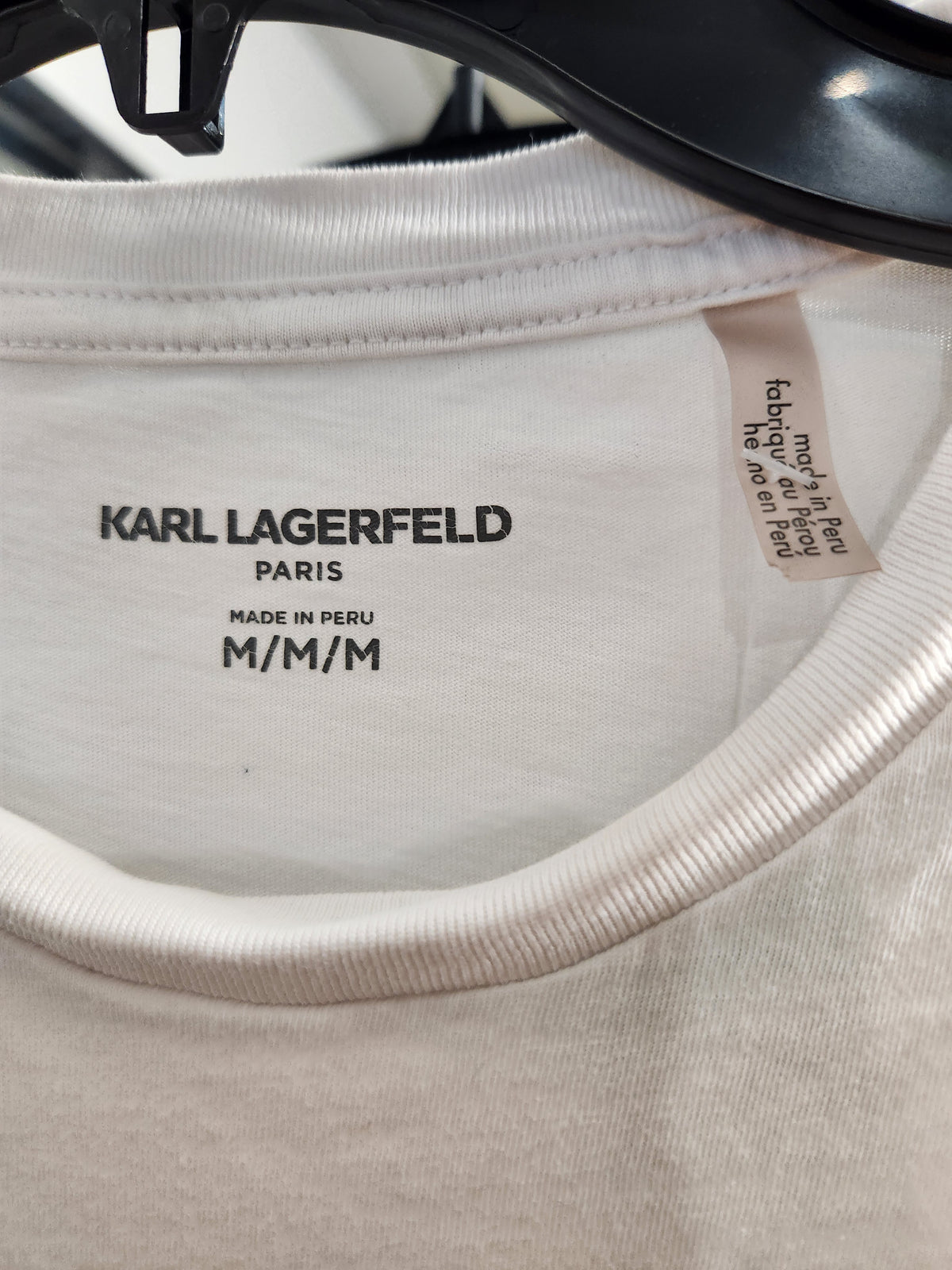 Close-up of the inside collar of a Karl Lagerfeld Paris T-shirt in a warm off-white or cream color, hanging on a black hanger. The brand name 'KARL LAGERFELD PARIS' is clearly printed on the inner neck label.