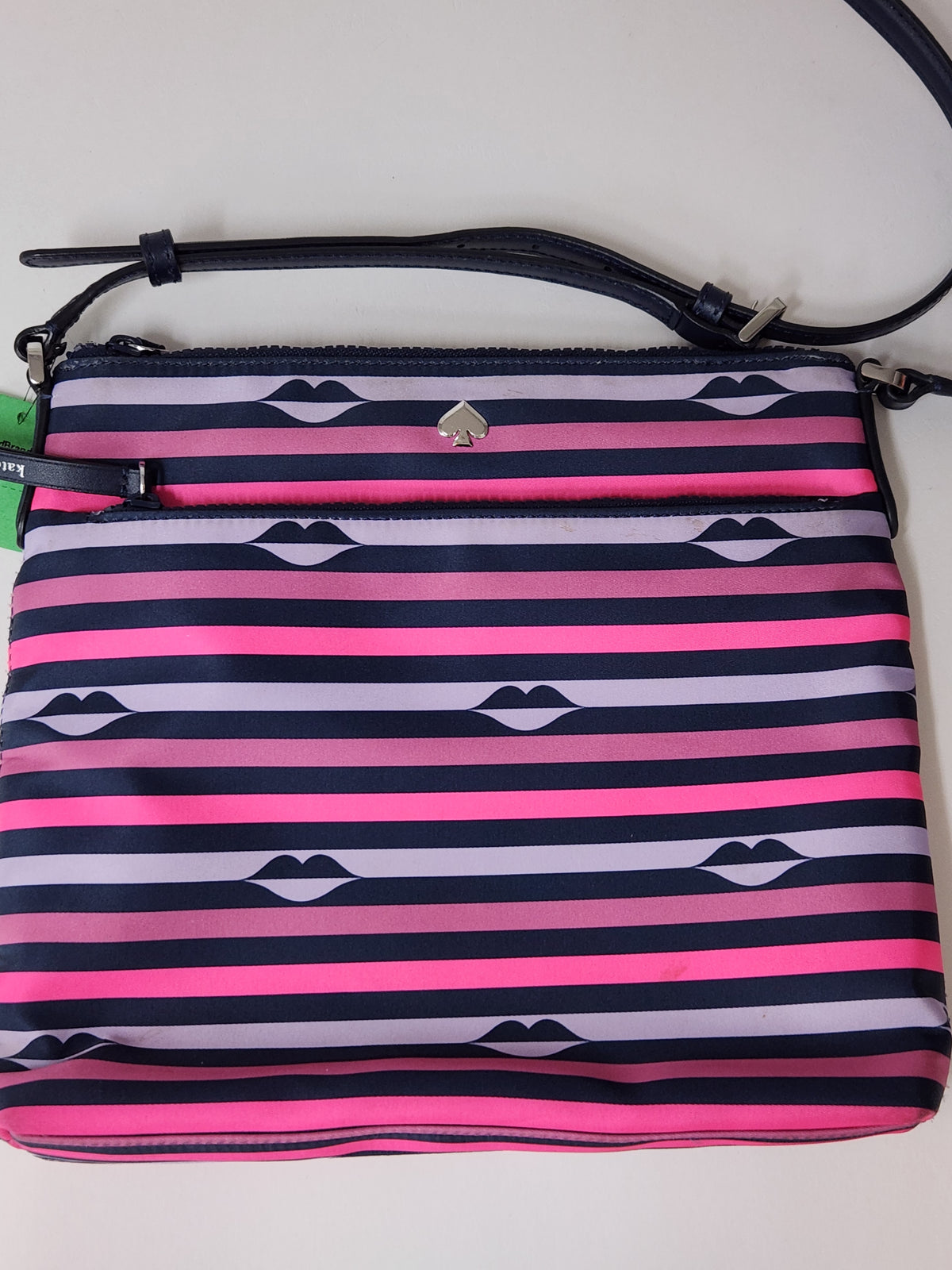 A striped crossbody bag with horizontal pink, purple, and navy blue stripes, featuring a repeating lip print motif and a silver-tone spade logo on the front pocket.
