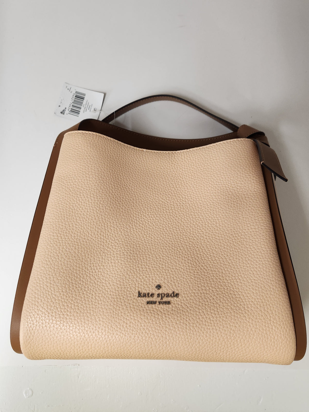 Kate Spade pebbled leather shoulder bag in beige and brown, featuring a top handle, a small top zip pocket, and the brand logo on the front, shown with a retail tag attached against a plain white background.